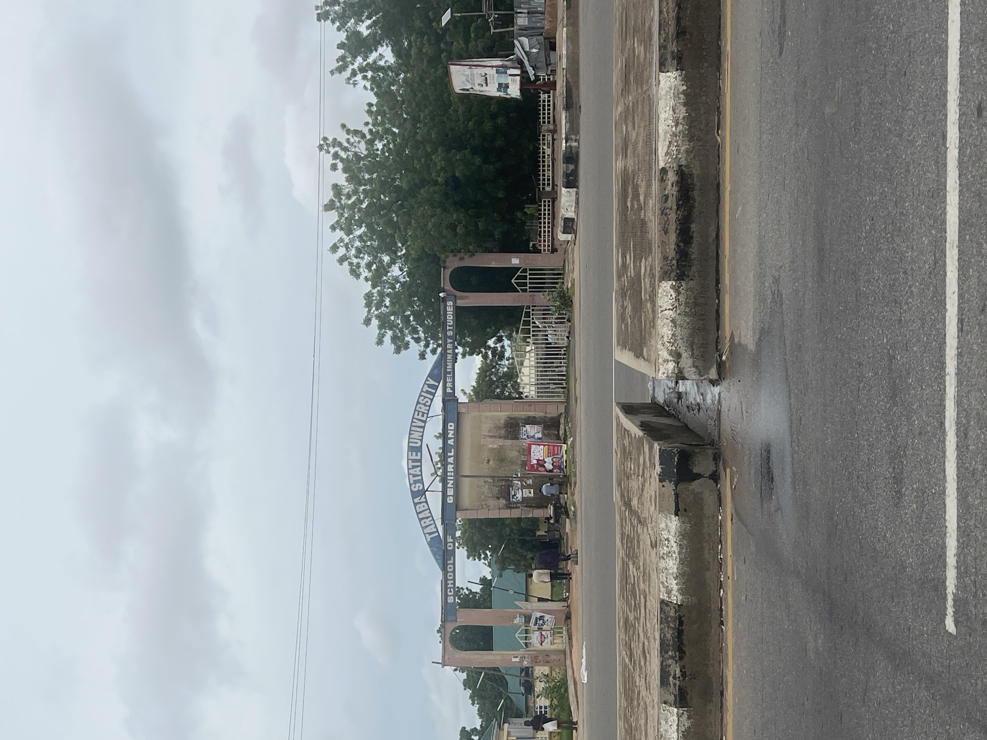 The image shows the entrance gate to Taraba State University, School of General and Preliminary Studies, located in Jalingo, Nigeria. The gate is a large archway with the university name prominently displayed. Several people are visible near the entrance, some walking and others standing. There are posters and advertisements on display at the entrance. The area appears to be a street in front of the university, with a road and concrete barriers. The sky is overcast, suggesting cloudy weather. Trees and buildings are visible in the background.