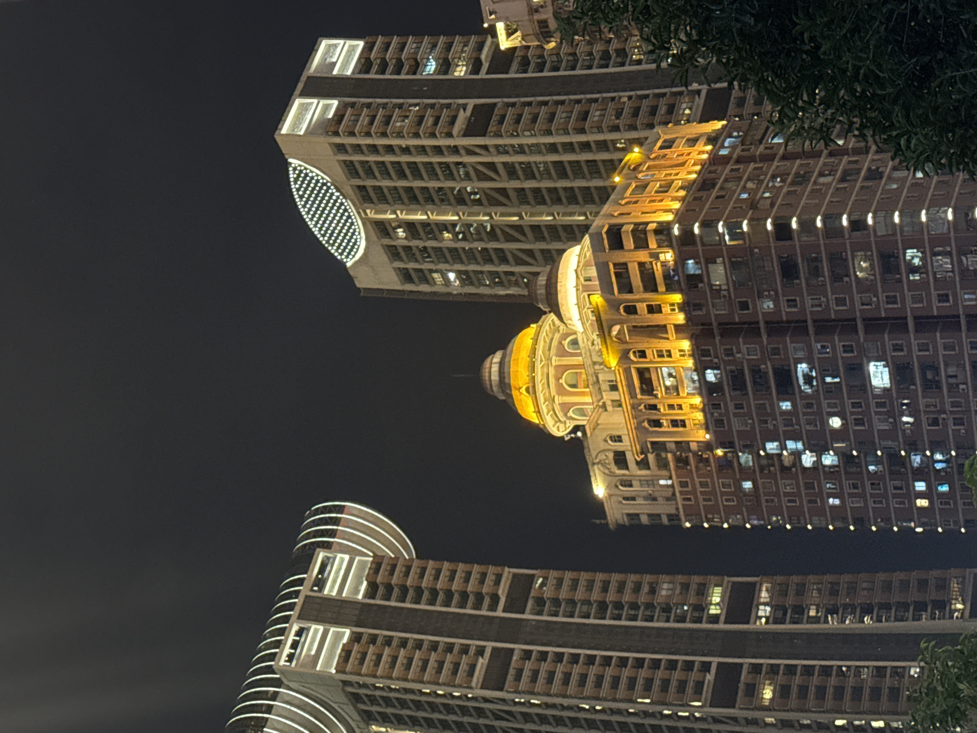 The image captures a nighttime view of several illuminated buildings in Yuzhong District, China. The primary focus is on a cluster of modern high-rise structures. The building on the far left is a tall, slender skyscraper with a distinctive curved top featuring white, illuminated concentric rings. Adjacent to it is a broader, taller building with a unique elliptical pattern of white lights on its upper section. In the center, a lower, ornate building features a prominent golden dome and is brightly lit with warm yellow lights along its upper floors, highlighting its architectural details. Further to the right, another tall building is partially visible, also lit from within and externally. The overall scene is dominated by the contrasting dark night sky and the vibrant artificial lighting of the urban architecture. There are no people or animals visible in the frame, and the image appears to be a static, architectural study.