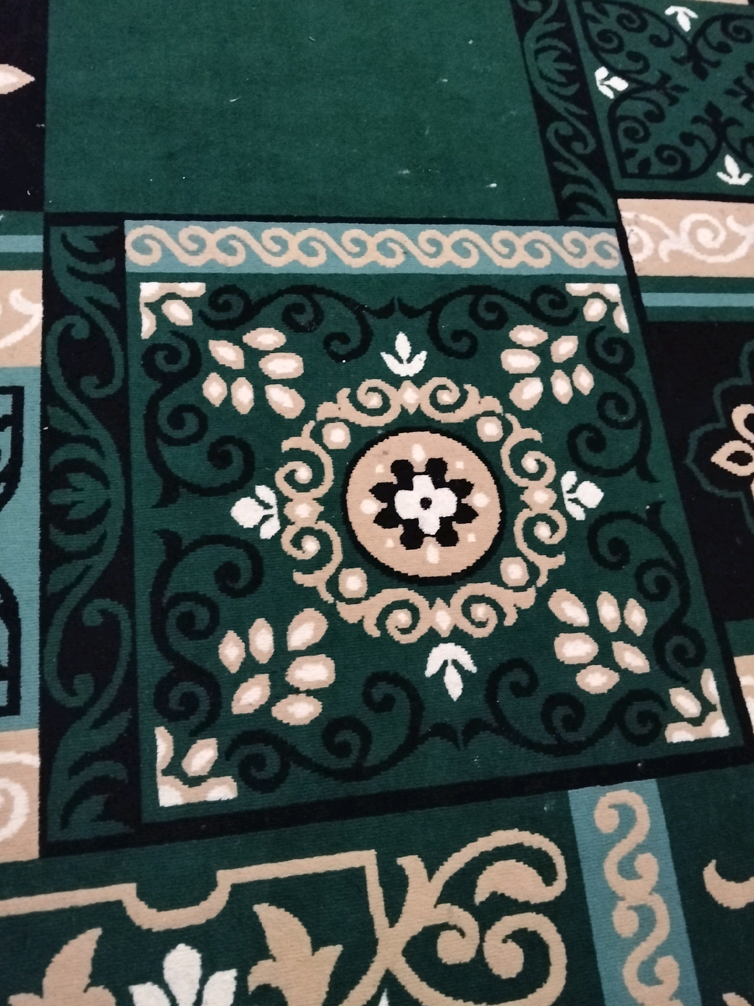 This is a close-up, overhead shot of a patterned green carpet, likely a prayer mat given the geometric and floral motifs. The carpet features a repeating design of dark green, black, beige, and white elements. The central focus is a square panel with intricate swirling patterns in black and beige, enclosing a circular motif with a stylized flower in black and white at its center, surrounded by a beige ring. Other parts of the carpet show broader bands with wave-like patterns in beige and light blue, and larger floral or scroll-like designs in beige against a green background. The texture appears to be that of a tufted carpet. The lighting suggests an indoor environment. The location provided, Suko Awin Jaya, Indonesia, is a place where such carpets might be found in homes, mosques, or community centers. There are no people or specific actions depicted in the image.
