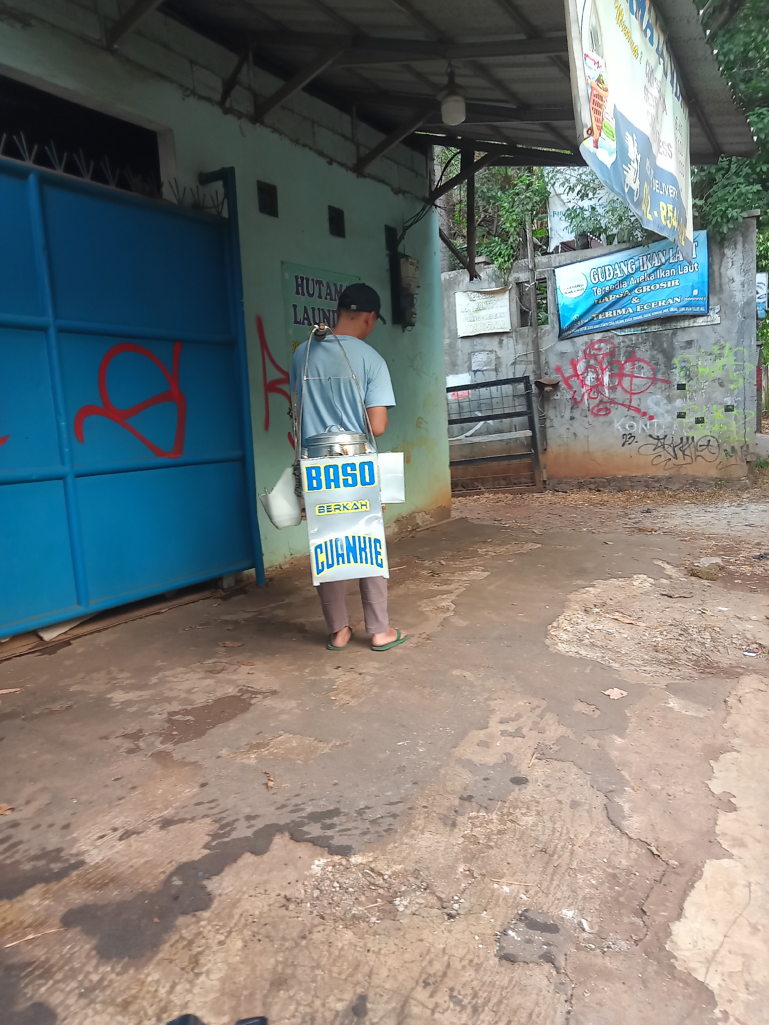 A rear view of a person wearing a light blue t-shirt and brown pants, carrying a food cart. The food cart has a metal container on top and two large rectangular signs hanging from its sides. The signs display "BASO BERKAH" and "CUANKIE" in large, bold lettering. The person is standing on a cracked, wet concrete ground. Behind the person, there is a blue metal gate with red graffiti. To the right of the gate, a light green building has a sign that reads "HUTAM LAUNDRY". Further to the right, a concrete wall is covered in graffiti, and a blue banner for "GUDANG IKAN LAUT" (Seafood Warehouse) is displayed. The environment appears to be an outdoor setting within a city, possibly in South Tangerang, Indonesia, given the signage.