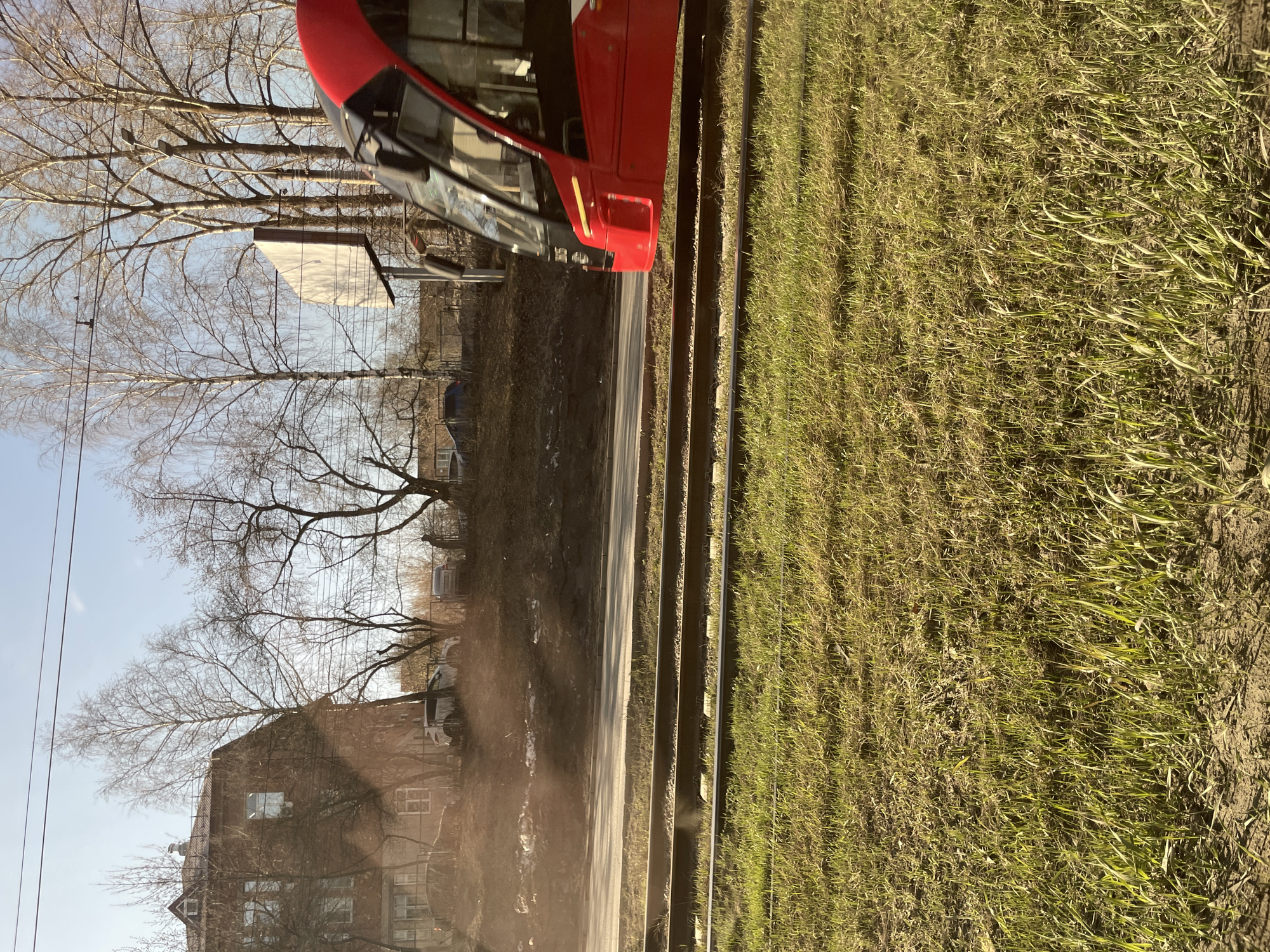 The image shows a street scene in Izhevsk, Russia. A red tram is partially visible on the right side of the frame. A road runs alongside the tram tracks. A grassy area is adjacent to the tracks. Across the road, there are buildings and trees without leaves. The sky is pale blue. Dust is present in the air near the lower part of the frame.
