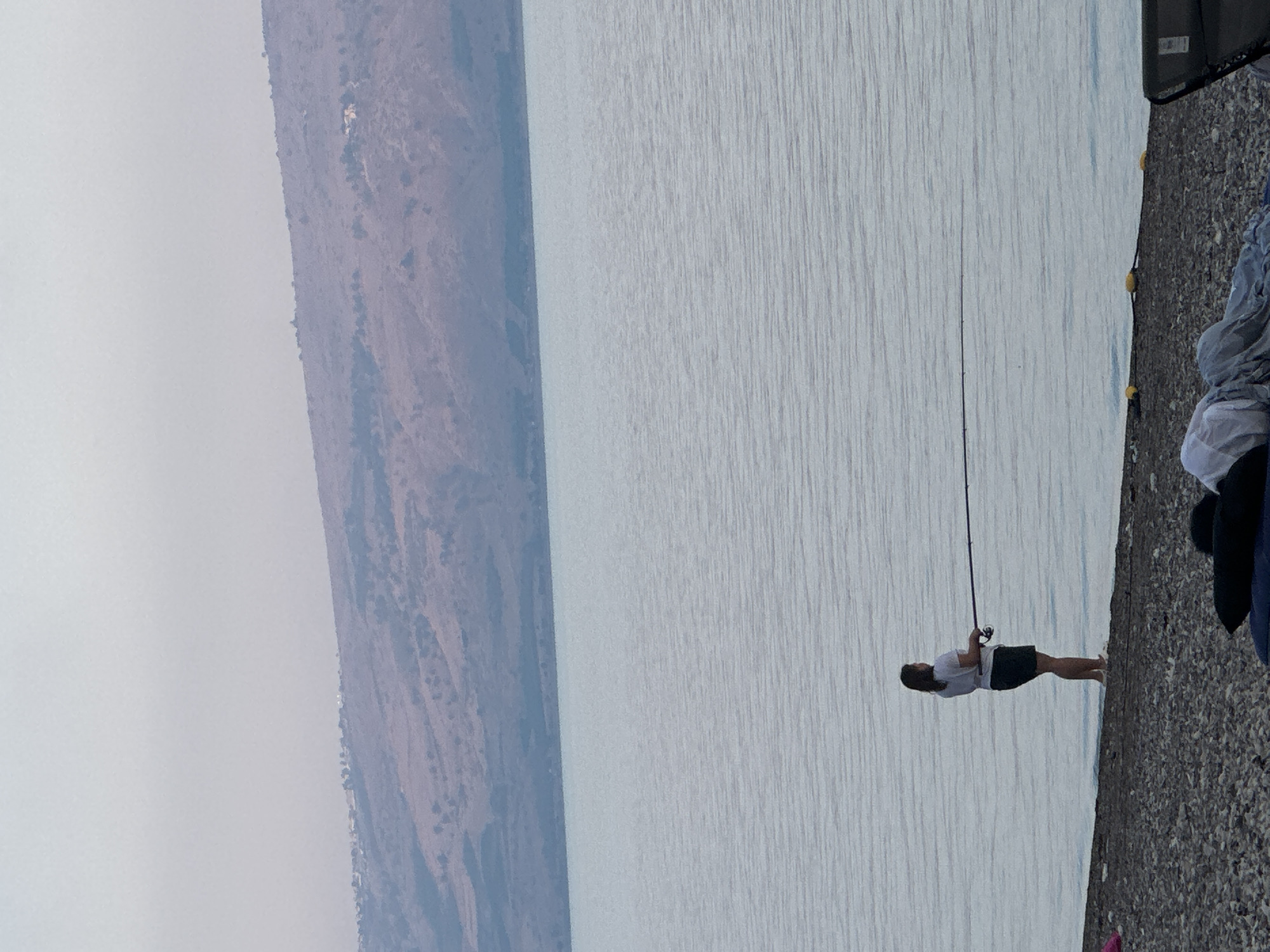 A woman stands on a pebble beach, facing the water and holding a fishing rod. She is wearing a white t-shirt and dark shorts. The water is calm and reflects the pale sky. In the background, a dry, hilly landscape stretches across the horizon, dotted with sparse vegetation and a few structures. The scene appears to be in Emek HaYarden Regional Council, Israel, likely near the Sea of Galilee. There are some scattered objects on the beach in the foreground, including cushions and folded fabric.