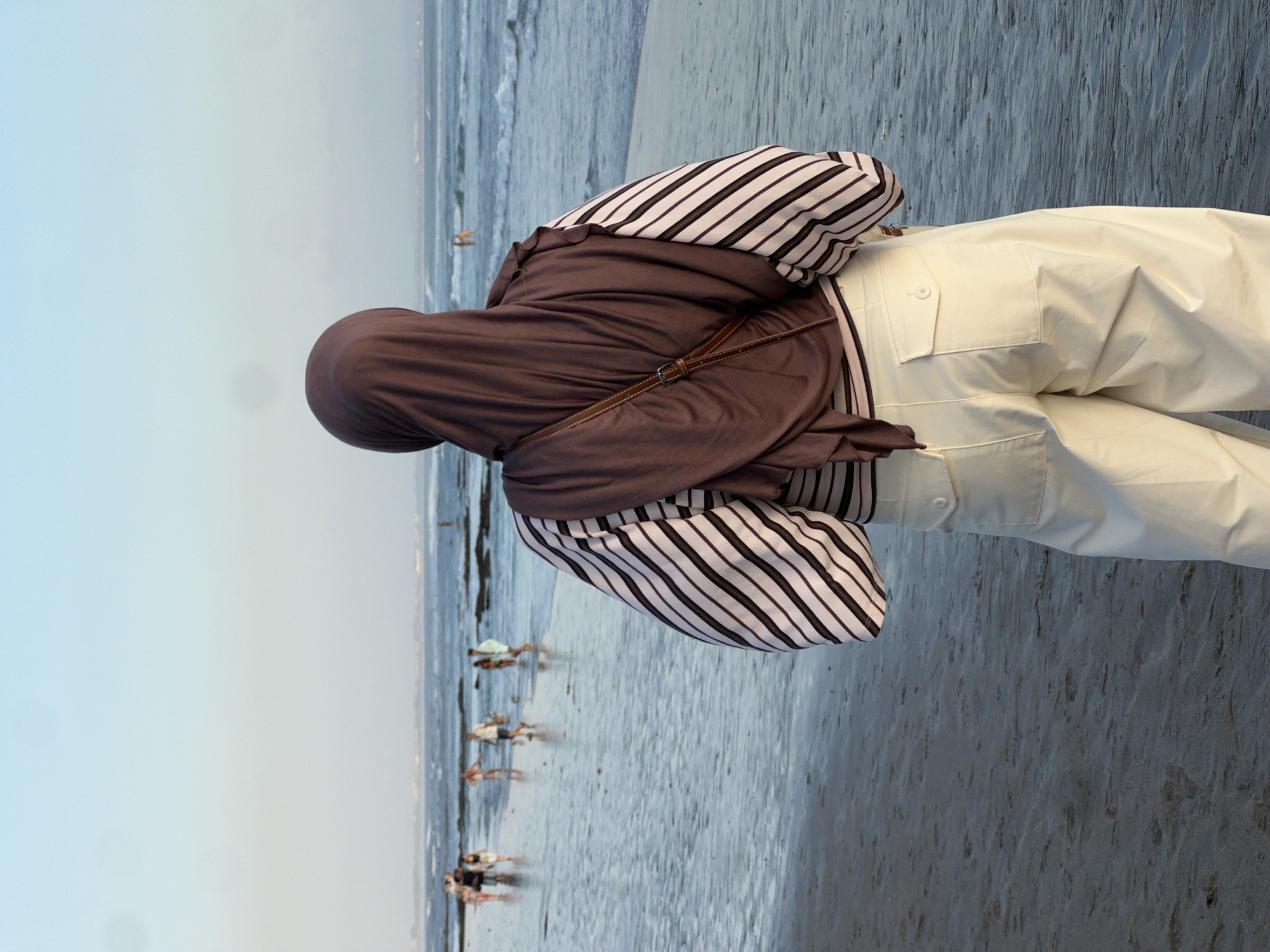 A person wearing a brown hijab and a striped shirt stands on a sandy beach, looking out at the ocean in North Kuta, Indonesia. The person's back is to the camera. The sky is clear and blue, with the sun setting in the distance. The ocean is calm, with small waves breaking on the shore. There are a few other people on the beach in the background, walking or standing near the water. The sand is wet and reflective, showing the footprints of passersby.