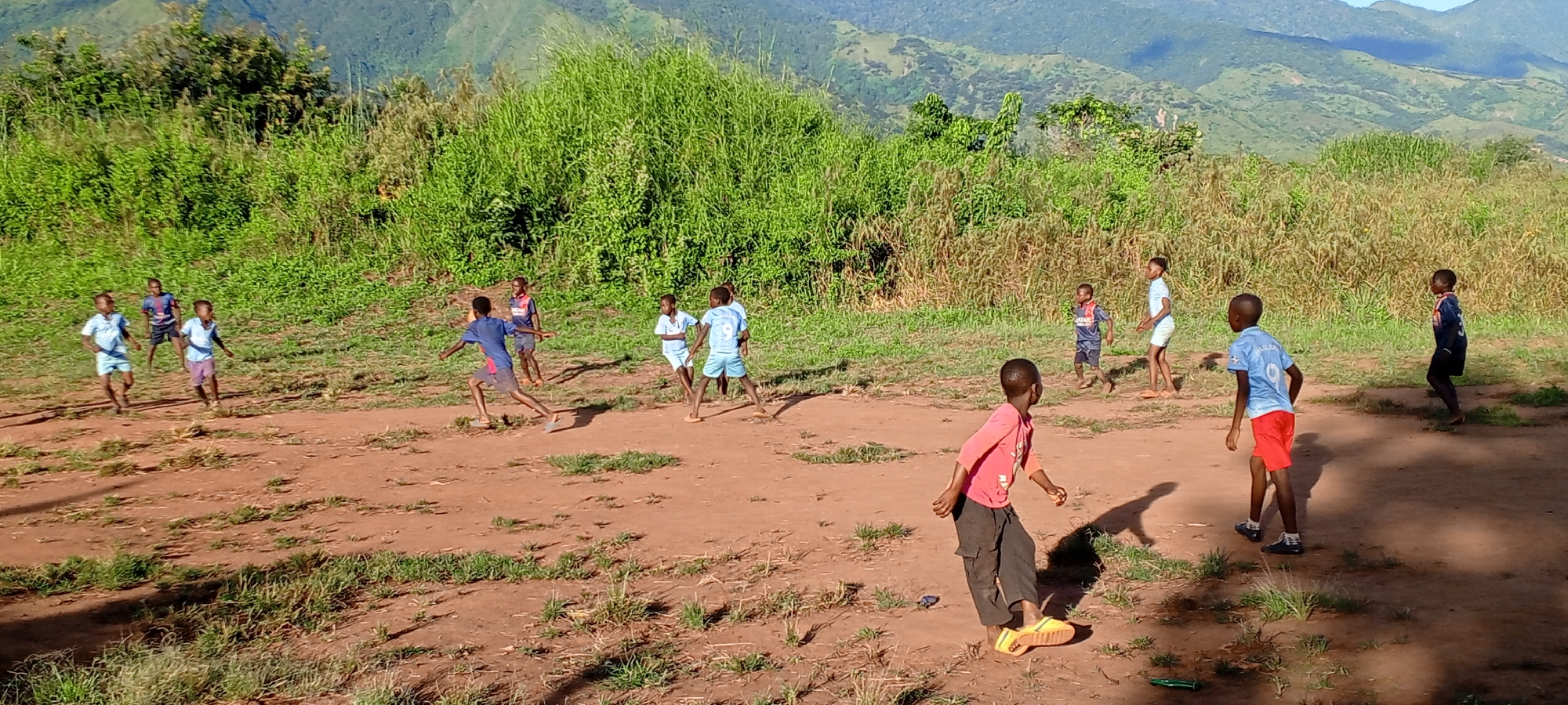 In an outdoor scene in Ngalula, Democratic Republic of the Congo, a group of approximately twelve children are engaged in a casual game. The children are dressed in a variety of casual clothing, including shorts, T-shirts, and sandals or bare feet. Some are wearing clothing with what appears to be soccer team branding. The setting is a dirt area interspersed with patches of grass and vegetation, suggesting an informal playing field. In the background, lush green hills and mountains rise in the distance. The sky is visible above the mountain range. The scene is bathed in natural daylight, with shadows cast by the children indicating the presence of sunlight. The scene conveys a sense of spontaneous outdoor activity.