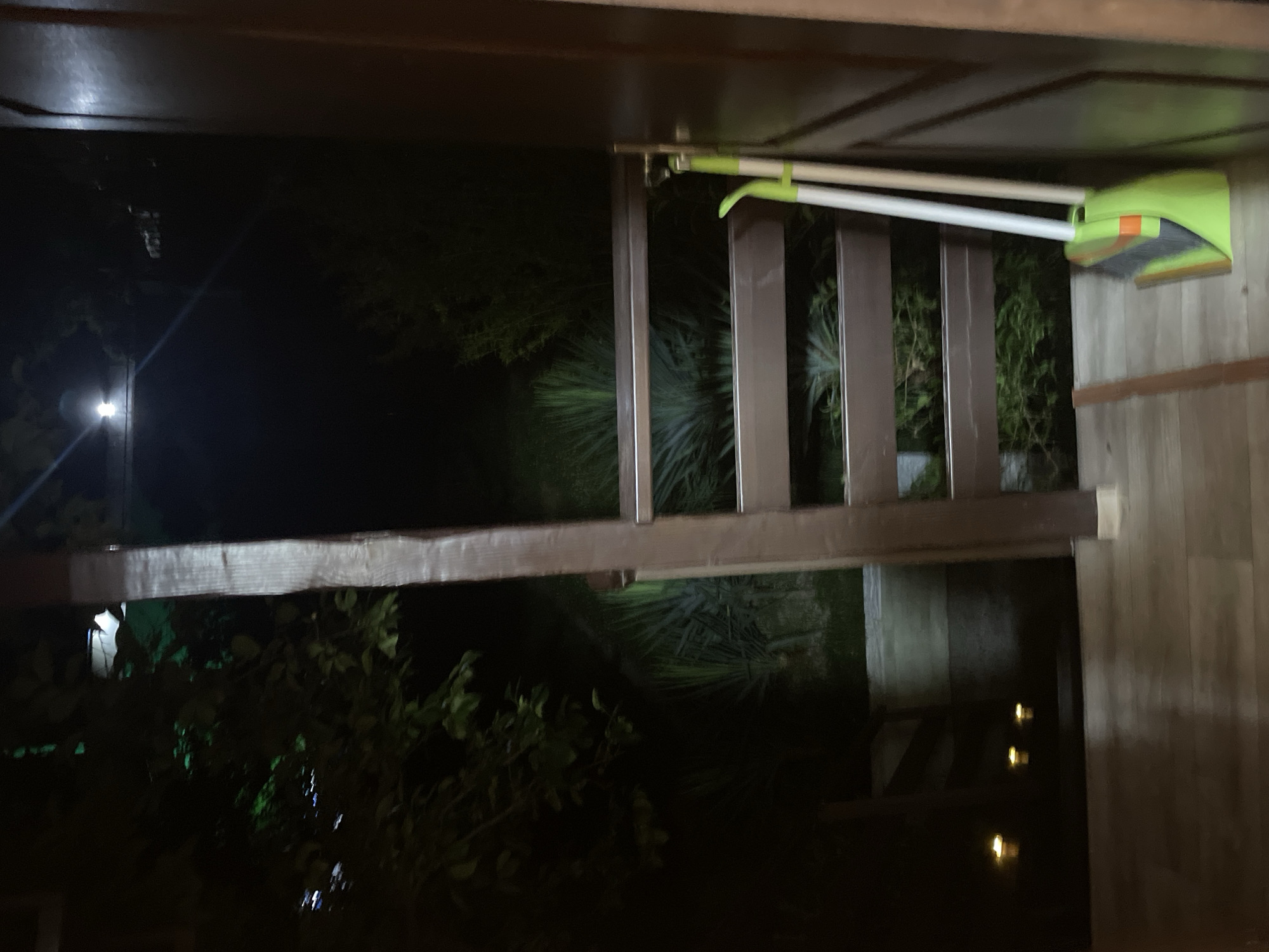 An outdoor scene at night, likely a porch or deck, is captured from a low angle. In the foreground, a wooden floor extends into the frame, with faint light reflecting off its surface. To the right, a closed wooden door stands vertically. Leaning against the door is a green and orange dustpan with white broom handles. A wooden railing with brown horizontal slats is visible behind the broom, with lush green foliage and some palm-like plants visible through the railing. In the background, a distant light source emits a bright glow, and several small, yellow lights are spaced along the floor to the left. The overall lighting is dim, with the exception of these light sources.