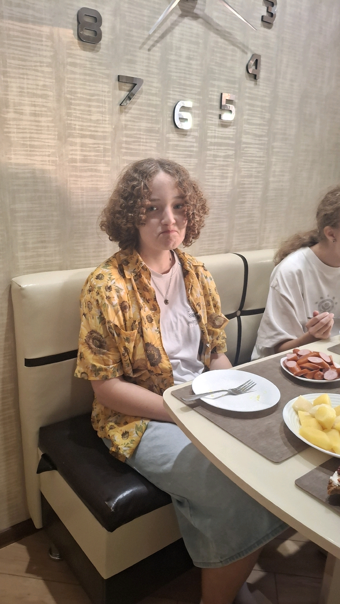 A medium shot shows a young person with curly brown hair, wearing a yellow sunflower-print shirt and light blue pants, seated on a booth at a dining table. Their expression is slightly sullen. To the right, another person's shoulder and arm are visible, also seated at the table. The table has plates of food, including sliced sausages and potatoes. In the background, a large wall clock with metallic numbers is visible. The overall setting appears to be an indoor dining area. The location is identified as Stowbtsy, Belarus.