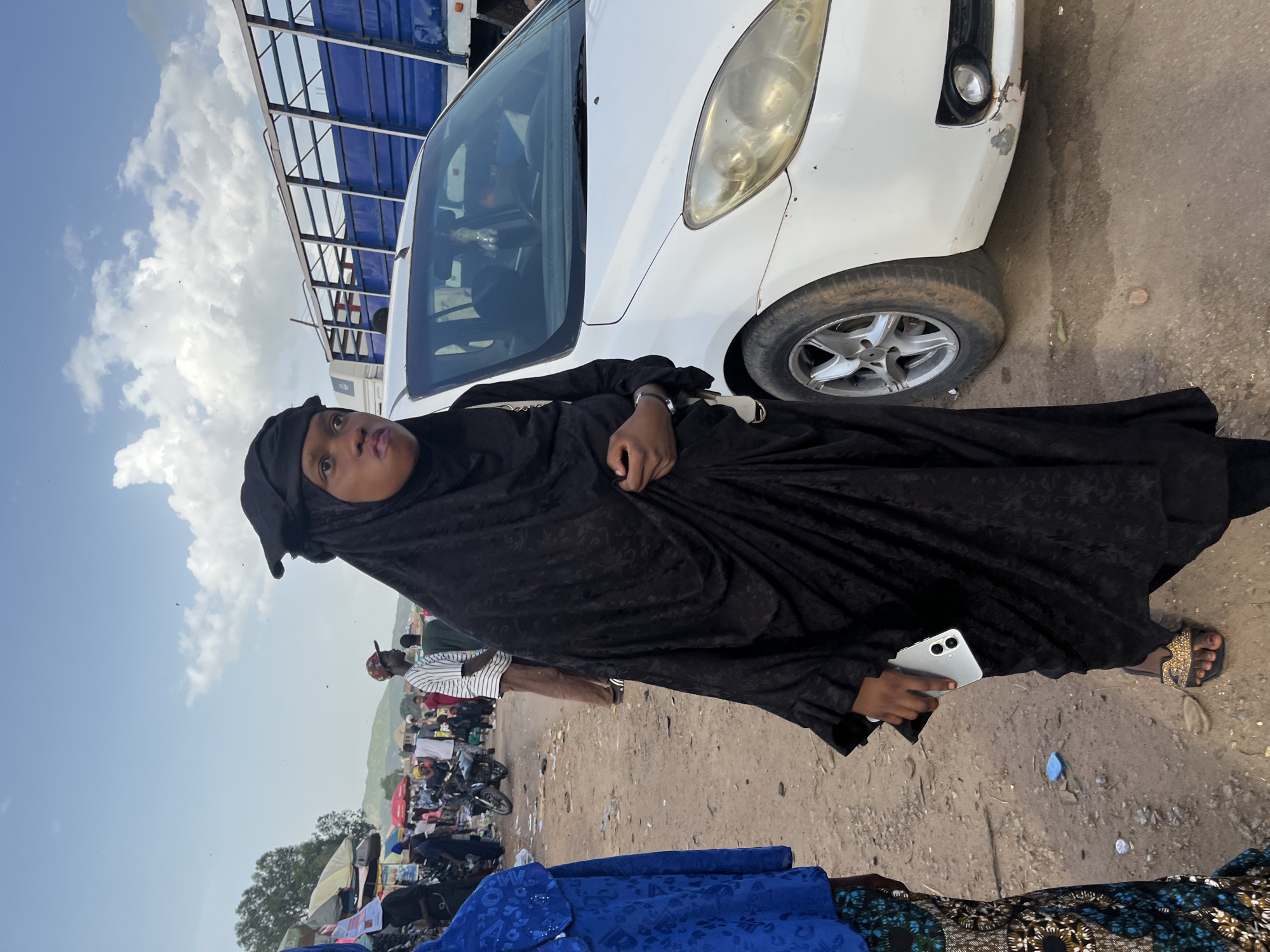 A young woman, wearing a black hijab and dress, stands in what appears to be a marketplace in Rafin Par, Nigeria. She is holding a silver smartphone in her right hand. To her left, another person is partially visible, wearing a blue top and a patterned skirt. Behind them, a white car is parked, and further in the background, a blue truck, several motorcycles, and other market stalls with people milling about are visible. The ground is dirt and somewhat littered. The sky is partly cloudy with a hazy sun.