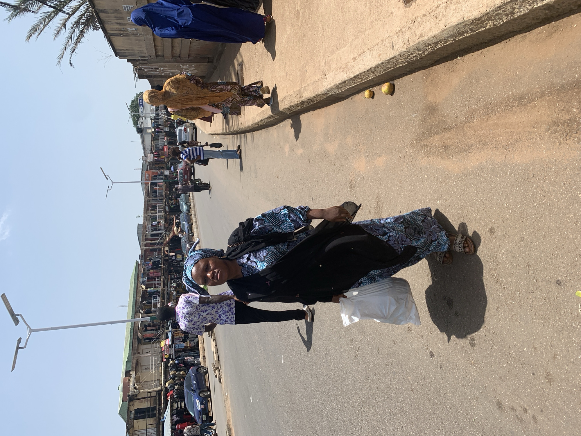 A woman wearing a blue and black patterned dress and a headscarf walks along a street in Jos, Nigeria. She carries a white plastic bag in her right hand. Another person wearing a purple and white patterned shirt is to her left. In the background, a busy market scene unfolds with shops lining the street and various vehicles, including cars and motorcycles, are visible. The sun is shining, casting shadows on the road, suggesting it is daytime. Several fruits, possibly mangoes, are scattered on the sidewalk. A lamppost stands on the left side of the road. The overall impression is a bustling street scene in a market environment.