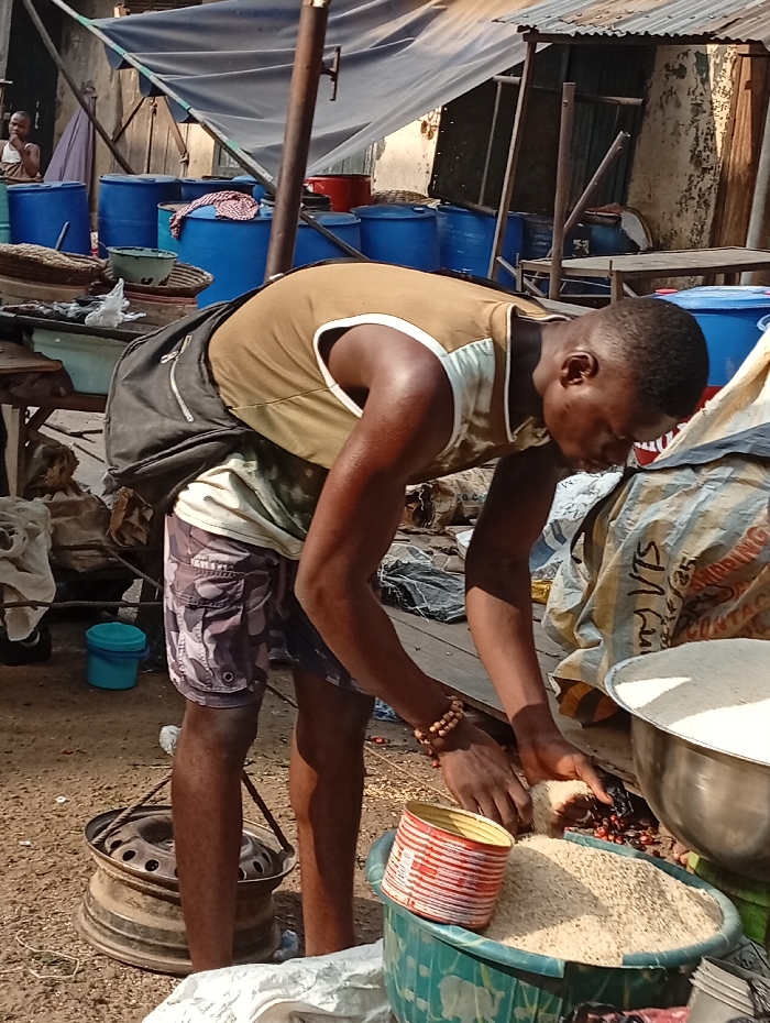 This image captures an outdoor scene, likely a market or a storage area, featuring a young man engaged in handling grains. The primary subject is a dark-skinned man, seen from the waist up as he bends forward, focused on the task at hand. He wears a light brown or khaki sleeveless top with white trim on the armholes and neckline, and camouflage-patterned shorts. A black backpack is slung over his back. On his right wrist, he wears a beaded wooden bracelet.
The man is actively scooping or sorting light-colored grains, likely rice, from a pile on the ground or within a green plastic basin. His hands are submerged in the grains, and a red and white striped metal can (possibly a repurposed food tin) sits next to the basin, suggesting he might be measuring or transferring the grains into it. Behind the green basin, a large silver metallic bowl also contains a significant amount of the same light-colored grains. Adjacent to the bowl is a large white sack or bag, on which fragmented text is partially visible, including what appears to be "SEA G...", "CINTA", "VLS", and some numbers like "25".
The background of the scene is filled with numerous blue plastic barrels of varying sizes, some with lids, suggesting storage or commercial activity. A light-colored tarp or canopy stretches overhead, supported by wooden or metal poles, providing shade. On the far left in the background, another person is partially visible, sitting next to one of the blue barrels. Other objects include a light blue plastic bucket on the ground, an old and rusty car wheel rim in the lower-left foreground, and a wooden table or stall structure with some items (possibly baskets or cloths) on it. Corrugated metal roofing is visible in the upper right.
The ground is uneven and appears to be dirt or sand, characteristic of an outdoor market or rural setting. The lighting suggests it is daytime, with bright sunlight and visible shadows. The overall impression is one of daily commerce or work in a bustling, informal environment.