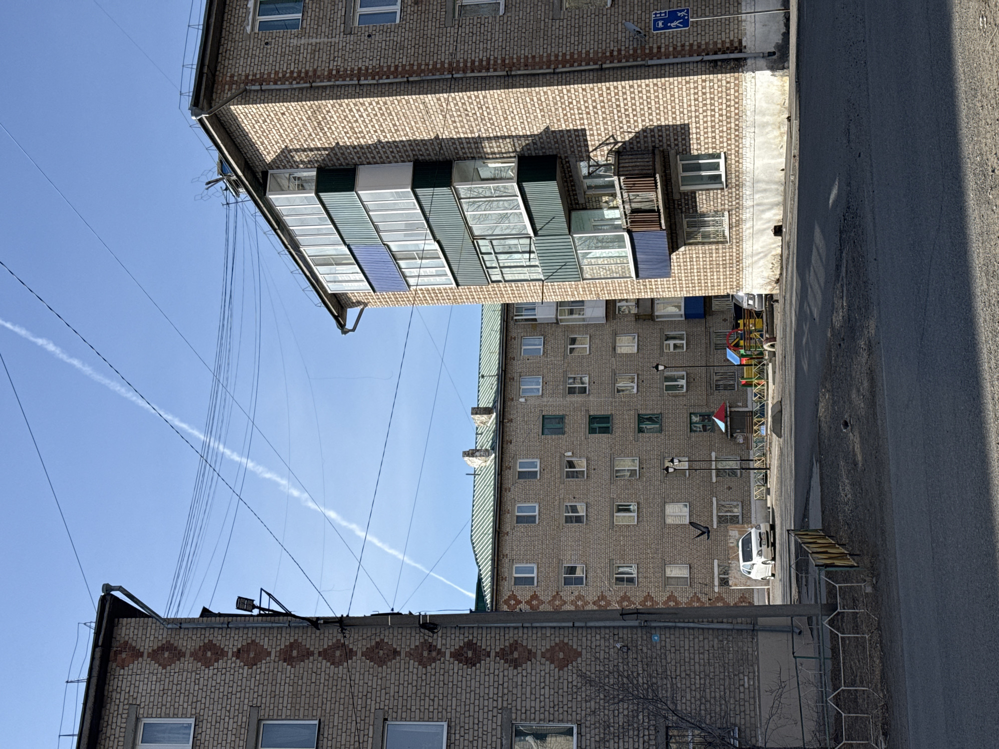 This image captures a quiet, sunny daytime scene in Borzya, Russia, depicting a residential street flanked by multi-story brick buildings. The architecture suggests a Soviet-era influence, with one prominent building featuring a large, multi-paneled glass-enclosed balcony or loggia structure. This building, along with others, is constructed from brick, some exhibiting decorative patterns with darker bricks.
The sky is a clear, bright blue, traversed by numerous overhead power lines and a distinct white airplane contrail. The bright sunlight casts visible shadows, indicating a mid-morning or afternoon setting.
At ground level, an asphalt road with a curb is visible. A white car is parked near a vibrant children's playground, equipped with colorful structures. A blue rectangular sign, mounted on a building wall, features graphic symbols for a bus stop and a pedestrian, indicating a public transport point. A metal fence with hexagonal elements runs alongside the road, and sparse tree branches, devoid of leaves, hint at late autumn, winter, or early spring. No people are visible, and the scene is devoid of overt activity, portraying an ordinary urban environment.