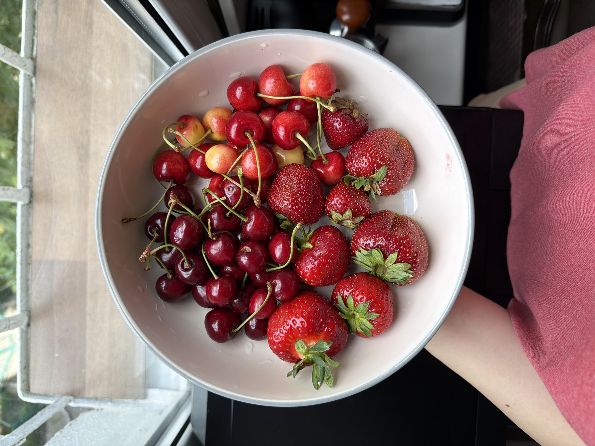 A top-down, medium shot captures a white bowl filled with ripe cherries and strawberries. The bowl rests on a dark surface, possibly a table or counter, in front of a window. Sunlight streams in, illuminating the fruits and casting subtle shadows. The cherries are a mix of deep red and lighter yellow-red hues, some with stems still attached. The strawberries are bright red, plump, and textured, with green leafy tops on some. A person's arm, with fair skin and a faint mole, is visible on the left side of the frame, holding the bowl. The person is wearing a reddish-pink garment. The background includes a glimpse of an outdoor scene through the window, with greenery and a white railing. The setting appears to be indoors, near a window, in a residential or kitchen environment.