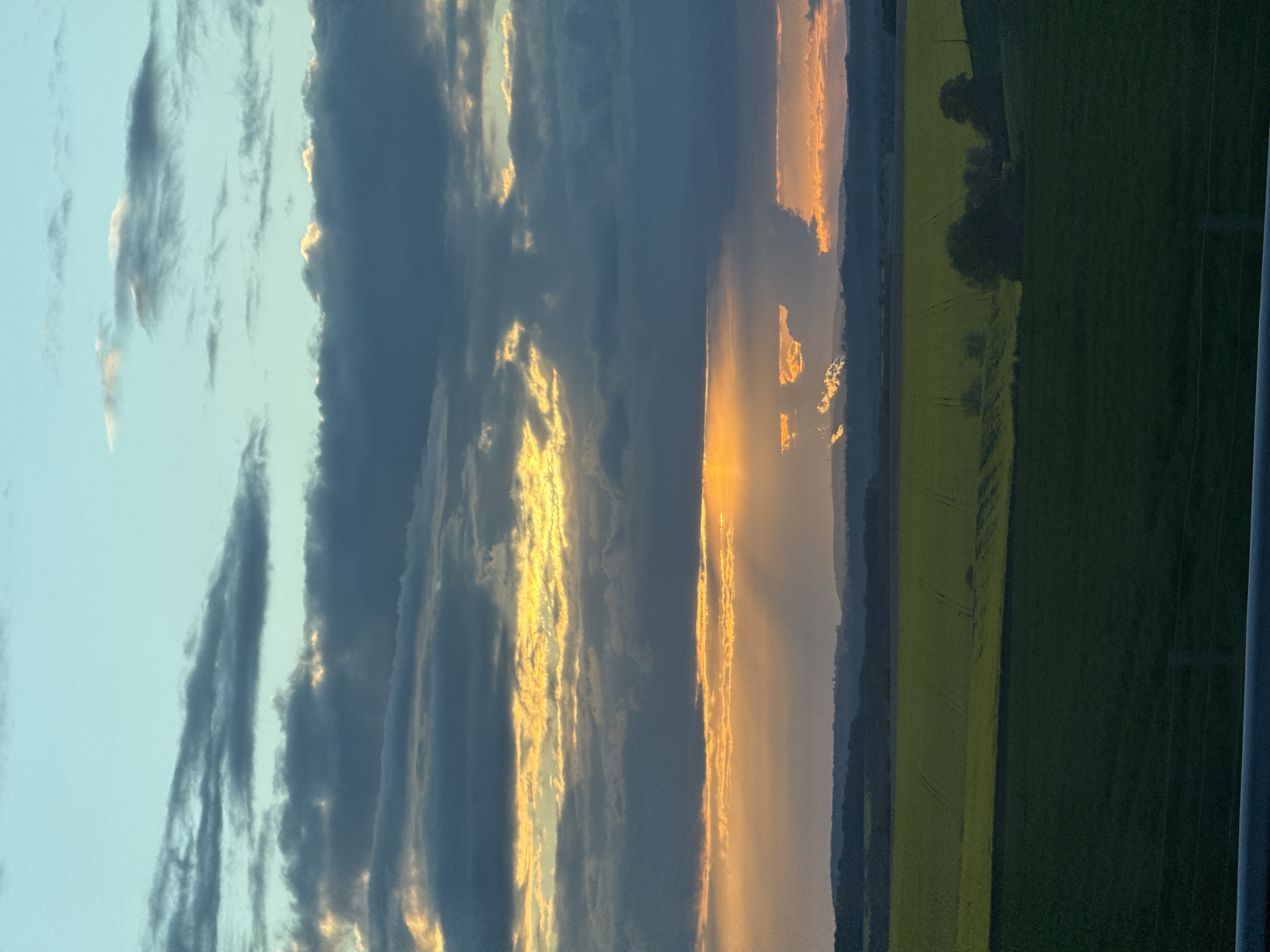 The image shows a landscape with a dramatic sky at sunset or sunrise. The sky takes up about half of the frame, featuring dark, heavy clouds with golden light breaking through. The light creates a radiant effect, particularly in the center of the frame. Below the sky, there's a land section with a field, appearing mostly green and yellow. The overall environment suggests a rural setting. It is located in the city of Jaignes, France.