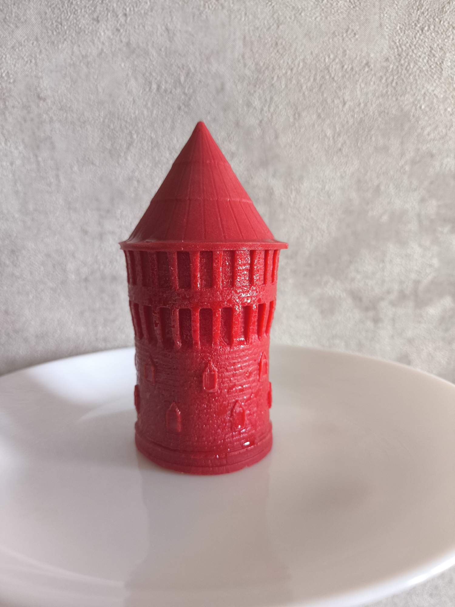 The image displays a vibrant red, miniature tower-shaped object standing centrally on a white, round plate. The tower features a conical roof with subtle horizontal ridges and a cylindrical body textured with intricate details resembling small, arched windows or decorative crenellations. The surface of the red object appears slightly glossy, suggesting a molded or 3D-printed material.
The white plate, serving as the base, exhibits a gentle sheen and reflects some ambient light. Behind the plate and tower, a light grey or off-white wall provides a soft, textured background. The overall lighting is diffused and even, indicating an indoor setting, possibly with natural or soft artificial light. No activity or event is taking place; the photograph presents a still-life composition. There are no people visible, nor is there any discernible text. The scene is situated in Moscow, Russia.
