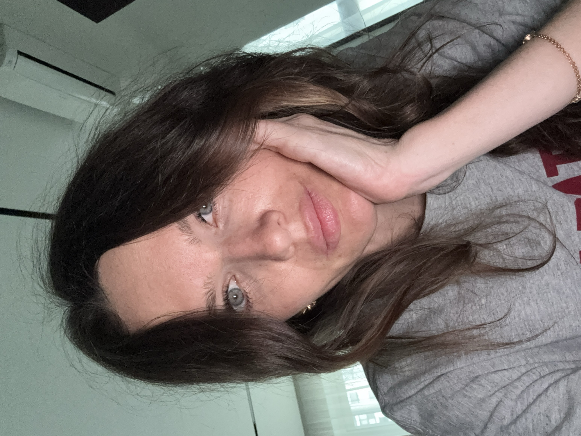 A selfie depicts a middle-aged woman with long brown hair and blue eyes, resting her face on her hand. She is wearing a gray t-shirt and a gold bracelet. The background shows a window with blinds and an air conditioning unit. The location in Khimki, Russia, is not evident from the visual information provided.