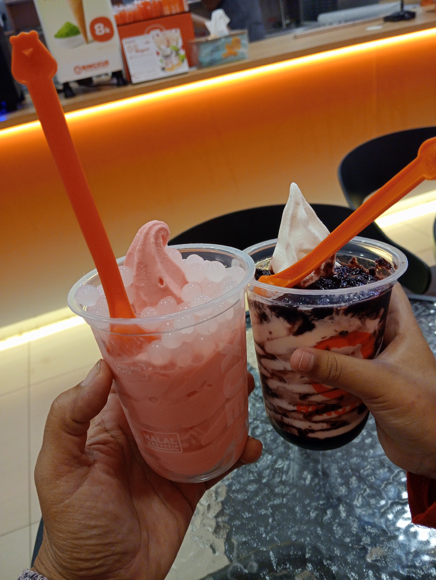 In this close-up, waist-level shot, two hands hold clear plastic cups filled with desserts, likely ice cream or a similar frozen treat, in what appears to be an indoor establishment. The lighting is warm, with an orange glow emanating from a strip light along a counter in the background. One cup contains a pink, soft-serve dessert with small, clear spheres, possibly boba or pearls, and is adorned with an orange spoon featuring a bear-shaped handle. The other cup holds a layered dessert with white and dark streaks, topped with a swirl of white soft-serve and a dark, possibly berry or chocolate, topping, also accompanied by an orange spoon. A blur of a counter with signage, including what looks like a calendar and branding, is visible behind the desserts. The table surface in the foreground has a speckled, reflective texture. The context of Pringsewu, Indonesia, suggests this scene likely takes place in a local cafe, dessert shop, or restaurant.