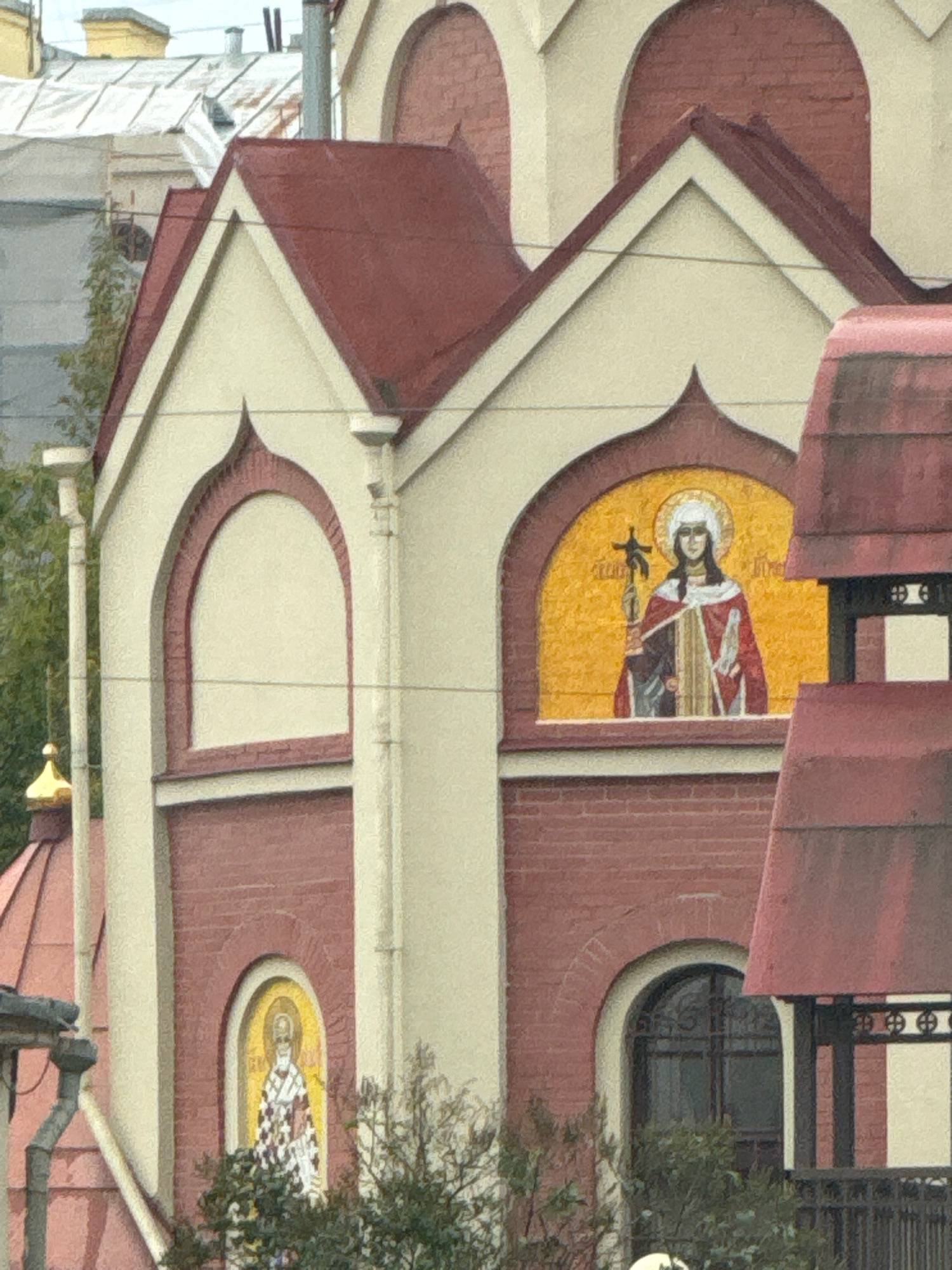 The image shows a close-up, eye-level view of the facade of a red brick building with cream-colored accents, likely an Orthodox church in Saint Petersburg, Russia. The architecture features arched windows and decorative mosaics depicting religious figures. The prominent mosaic in the upper center shows a female saint with a halo, holding a cross and dressed in red and gold robes. Below and to the left, a smaller mosaic depicts another saint in checkered robes. Red, pitched roofs with decorative finials are visible at various levels. The building is partially obscured by greenery and scaffolding on the right side. The sky is overcast.