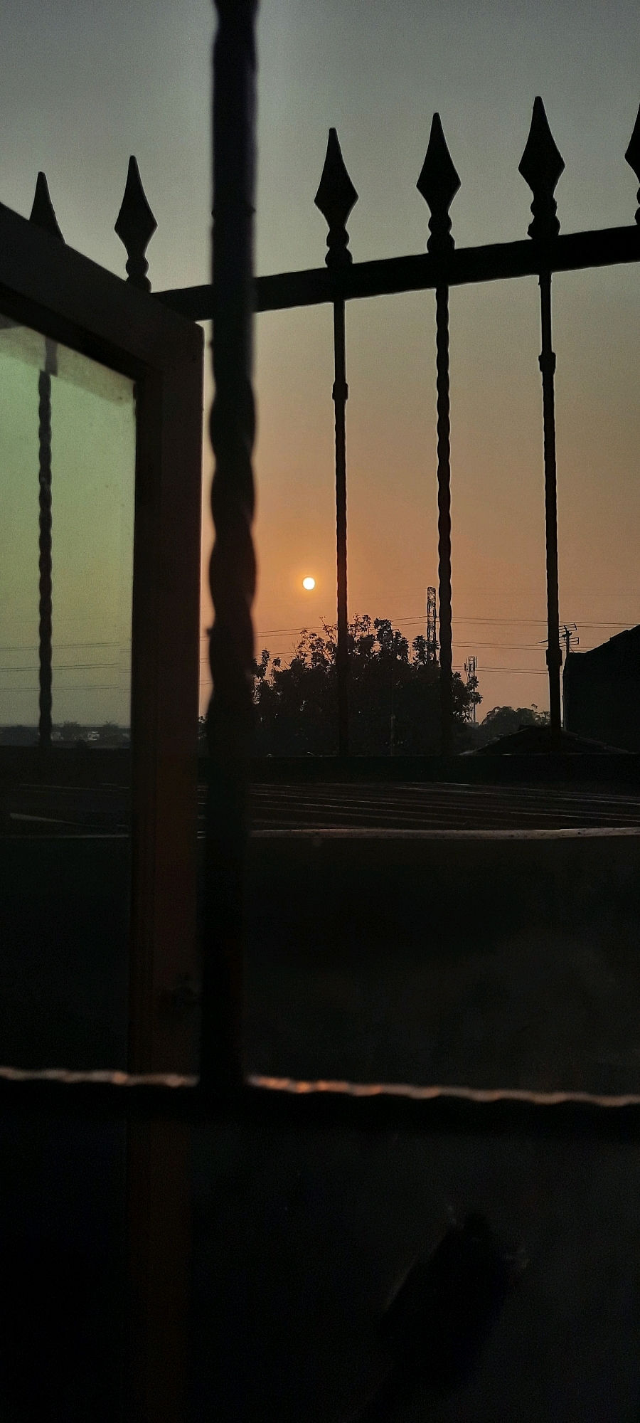 The image depicts a sunset viewed through a barred window. The sun, a small, bright orb, is visible in the hazy sky. Silhouetted against the sky are trees and what appear to be telecommunication towers. In the foreground, the dark bars of the window frame the scene, and on the left, a portion of a window pane reflects a greenish light. The overall impression is one of looking out from an enclosed space towards an urban landscape at dusk. The presence of telecommunication towers suggests an urban environment. While the location is identified as East Jakarta, Indonesia, this specific detail is not visually discernible from the image itself. The scene is static and does not show any people or active interactions.