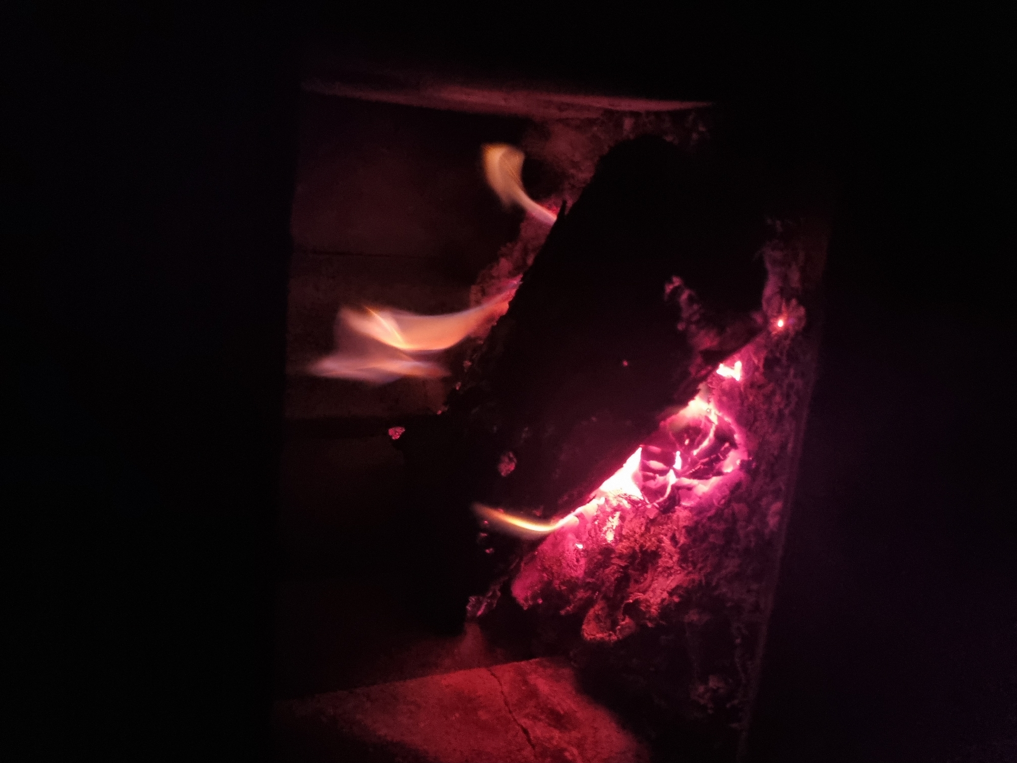 A wood-burning stove in Elena, Bulgaria is filled with burning logs. The logs appear charred and red with embers. Flames lick at the wood, with some escaping into the space above. The interior of the stove is dark and only partially visible. The rest of the surrounding room is in deep shadow.