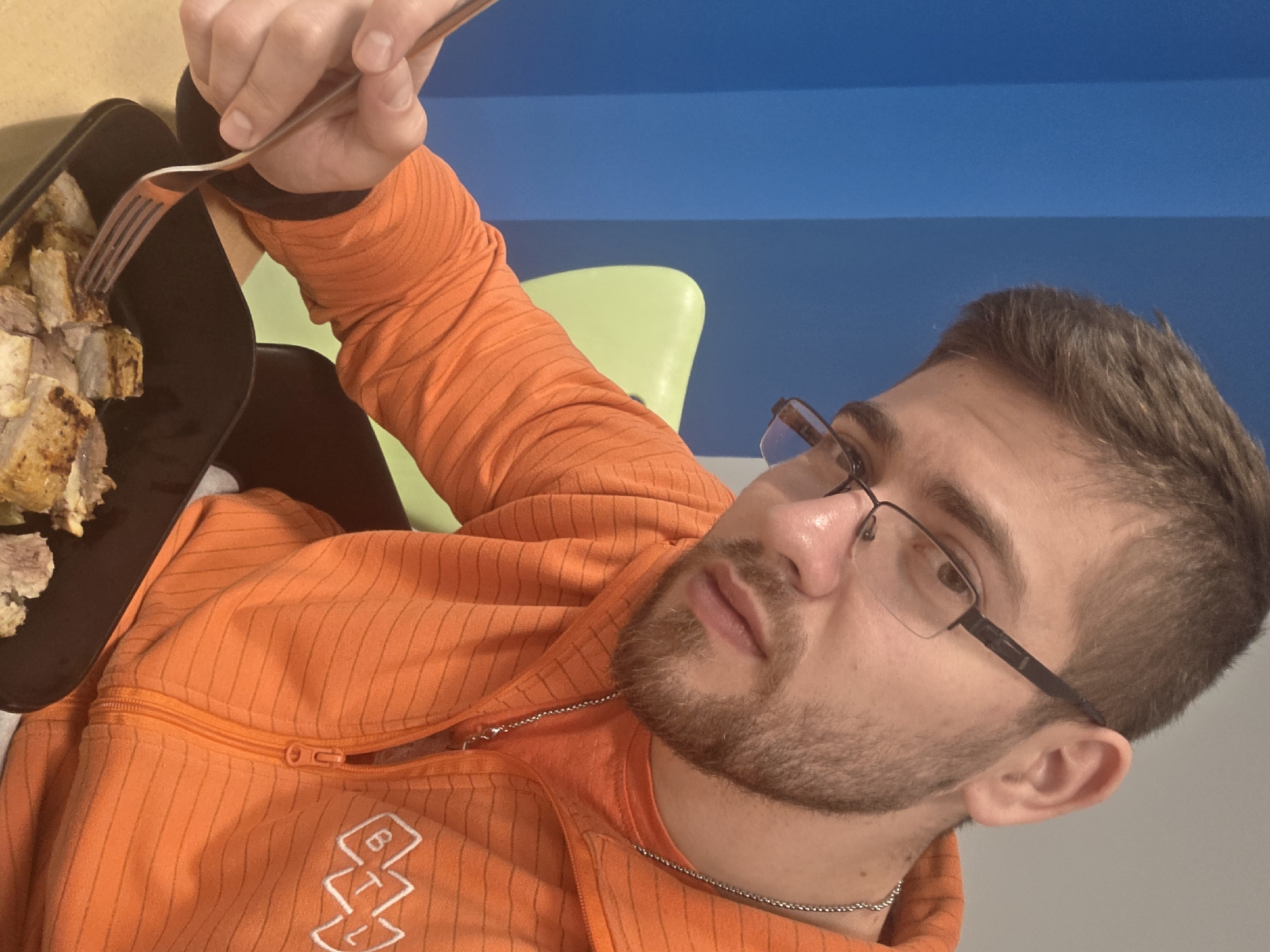 A man with short brown hair and a beard is seated, holding a fork and a black plate with cooked food. He is wearing glasses and an orange zipped jacket with a white logo. The background consists of a blue wall with a white stripe and a lime green chair. The photo was likely taken indoors. There is no information on this image that indicates the location is in the city of Plovdiv, Bulgaria.
