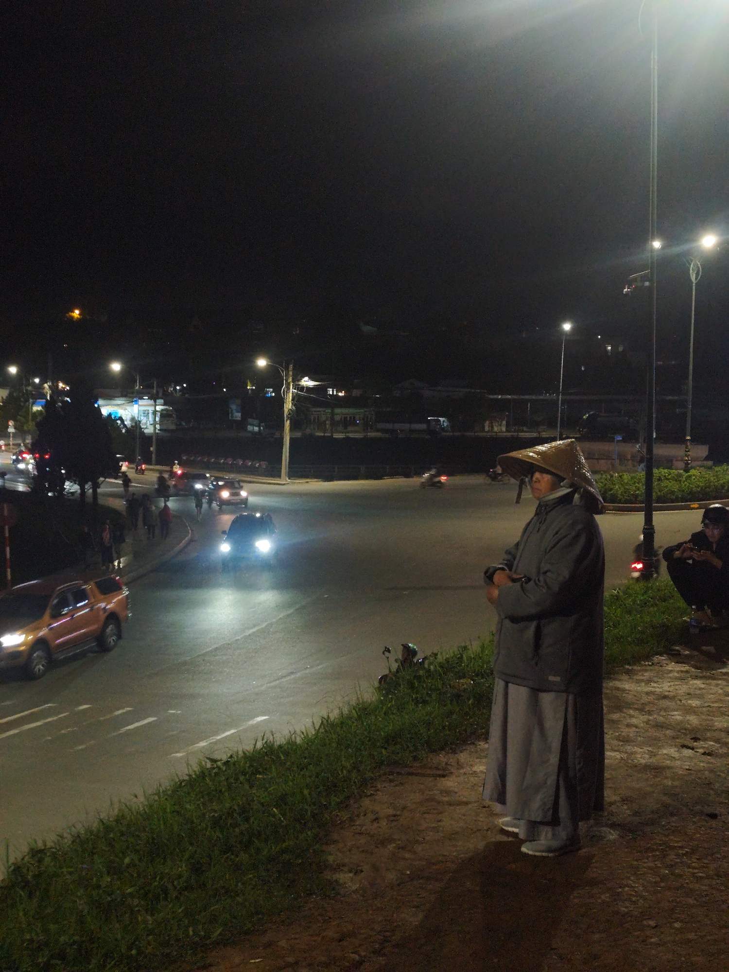 The photo was taken at night in Da Lat, Vietnam. At the edge of a road with a raised grass verge, a person wearing a gray coat, gray skirt, and a woven conical hat stands with arms crossed. To their right, a person sits crouched, facing away from the camera. In the background, a well-lit street intersection displays cars, motorcycles, and pedestrians. Street lights illuminate the road, and buildings are visible in the distance. The ground beside the grass verge is brown and appears damp.