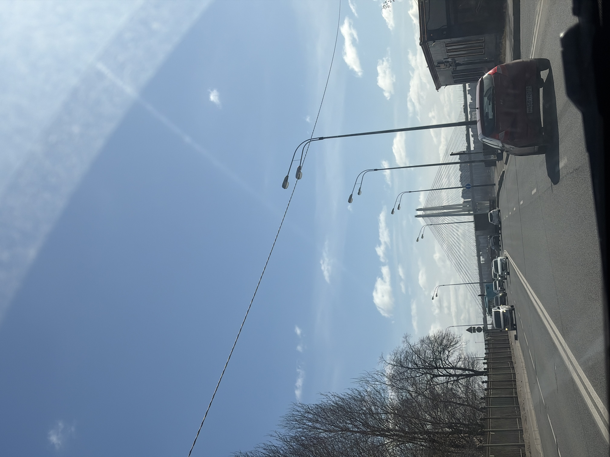 The image shows a view of a road with several cars and a bridge in the distance. The sky is blue with some clouds. The foreground includes a tree with bare branches and streetlights lining the road. In the distance, a large cable-stayed bridge can be seen. Based on the architecture of the bridge and the surrounding urban landscape, the location is likely Saint Petersburg, Russia, specifically the cable-stayed Bolshoy Obukhovsky Bridge over the Neva River. The photograph seems to have been taken from inside a moving vehicle.
