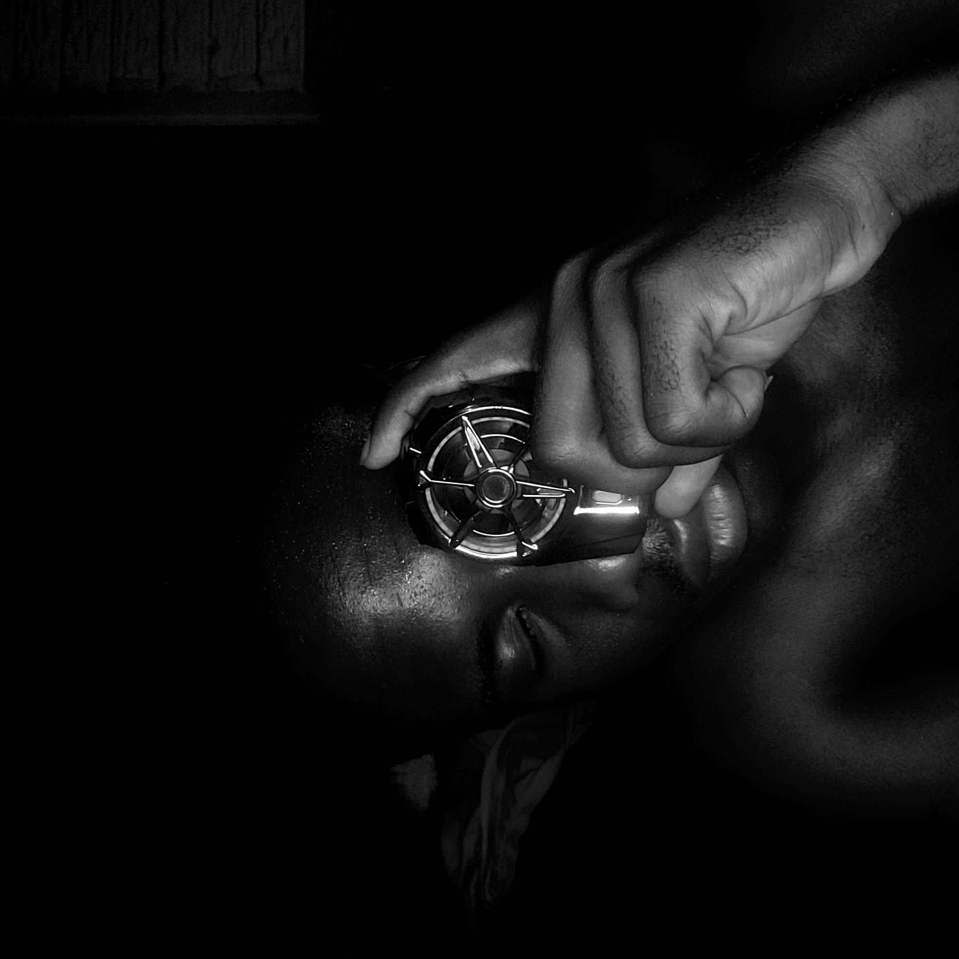 The image is a black and white close-up shot featuring a dark-skinned individual lying down with their eyes closed. A hand is holding a small, circular object with a metallic, fan-like structure against the individual's face, near their mouth. The surrounding environment is dark and largely undefined. Location cannot be confirmed.