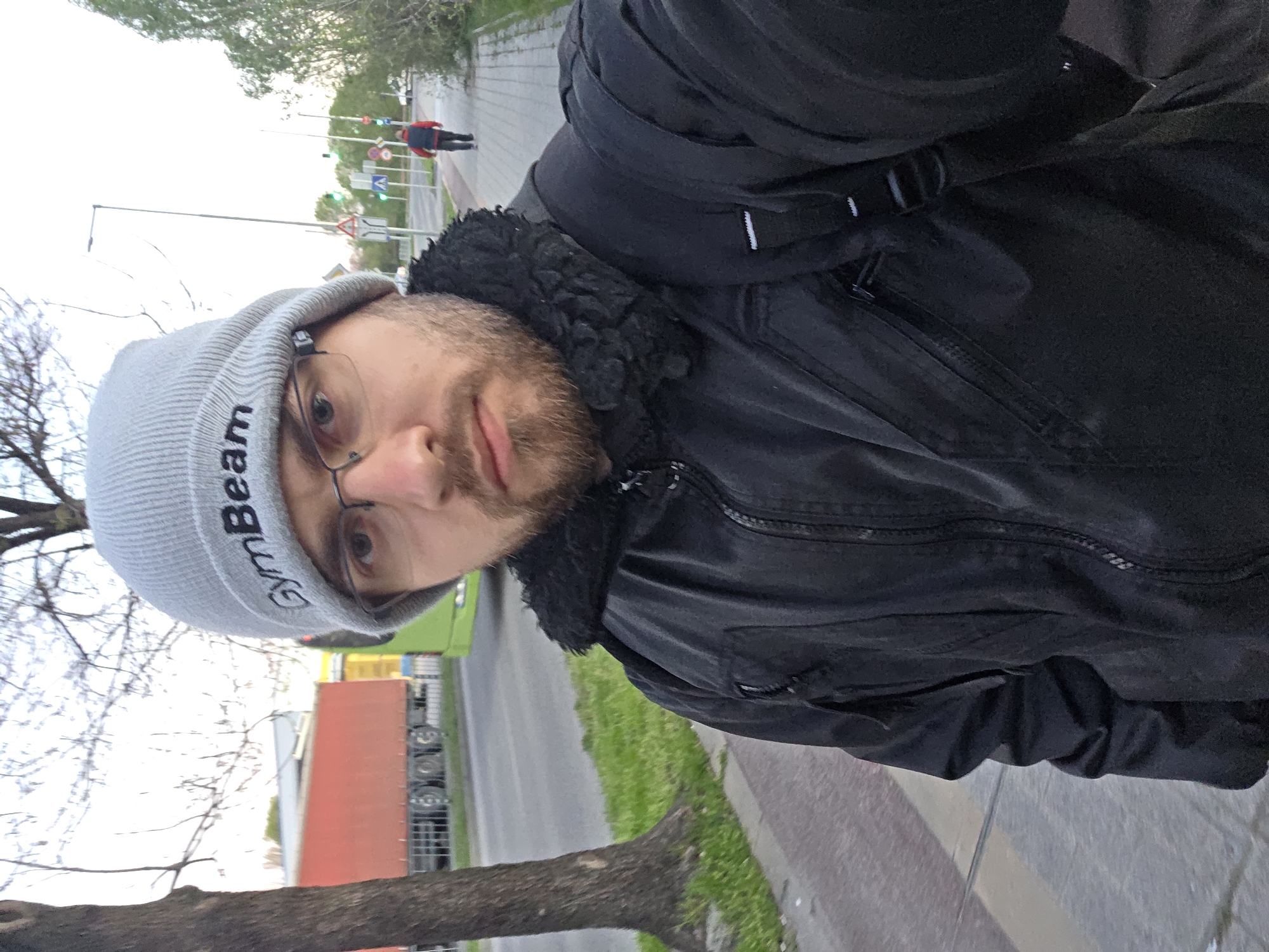 A man wearing a gray beanie with the word "GymBeam" vertically printed on it, black-rimmed glasses, and a black jacket with a fur collar takes a selfie. He has a beard and appears to be on a sidewalk. In the background, there is a street with a truck, some greenery, and a person walking further down the sidewalk. Based on the style of streetlights and road signs, and the general layout, it is likely the photo was taken in Plovdiv, Bulgaria.