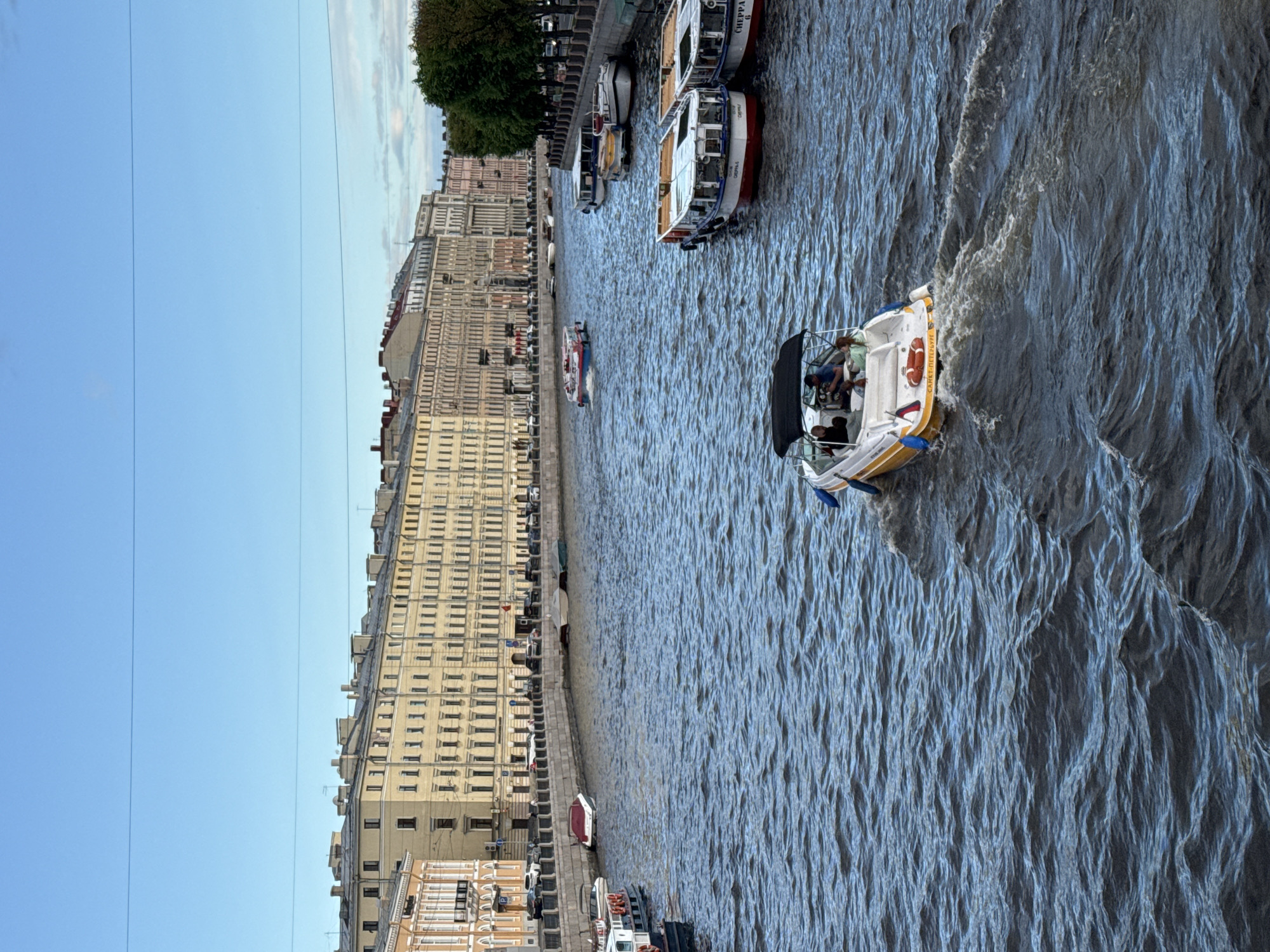 A clear, wide canal with blue water runs through a European city, bordered by light yellow and orange buildings with classical architecture. Several boats navigate the canal. In the foreground, a white and yellow motorboat with a black canopy is moving away from the camera, carrying at least three people, one of whom is steering. Further up the canal, a red and white tour boat with a flat roof is moving towards the camera. On the right bank, several tour boats are docked, including one with a red stripe and another with a white roof. On the left bank, more buildings line the canal, with cars parked along the street behind a stone embankment. The sky is a clear, light blue. The scene appears to be St. Petersburg, Russia, likely on one of its many canals or rivers, given the architecture and prevalence of tour boats.
