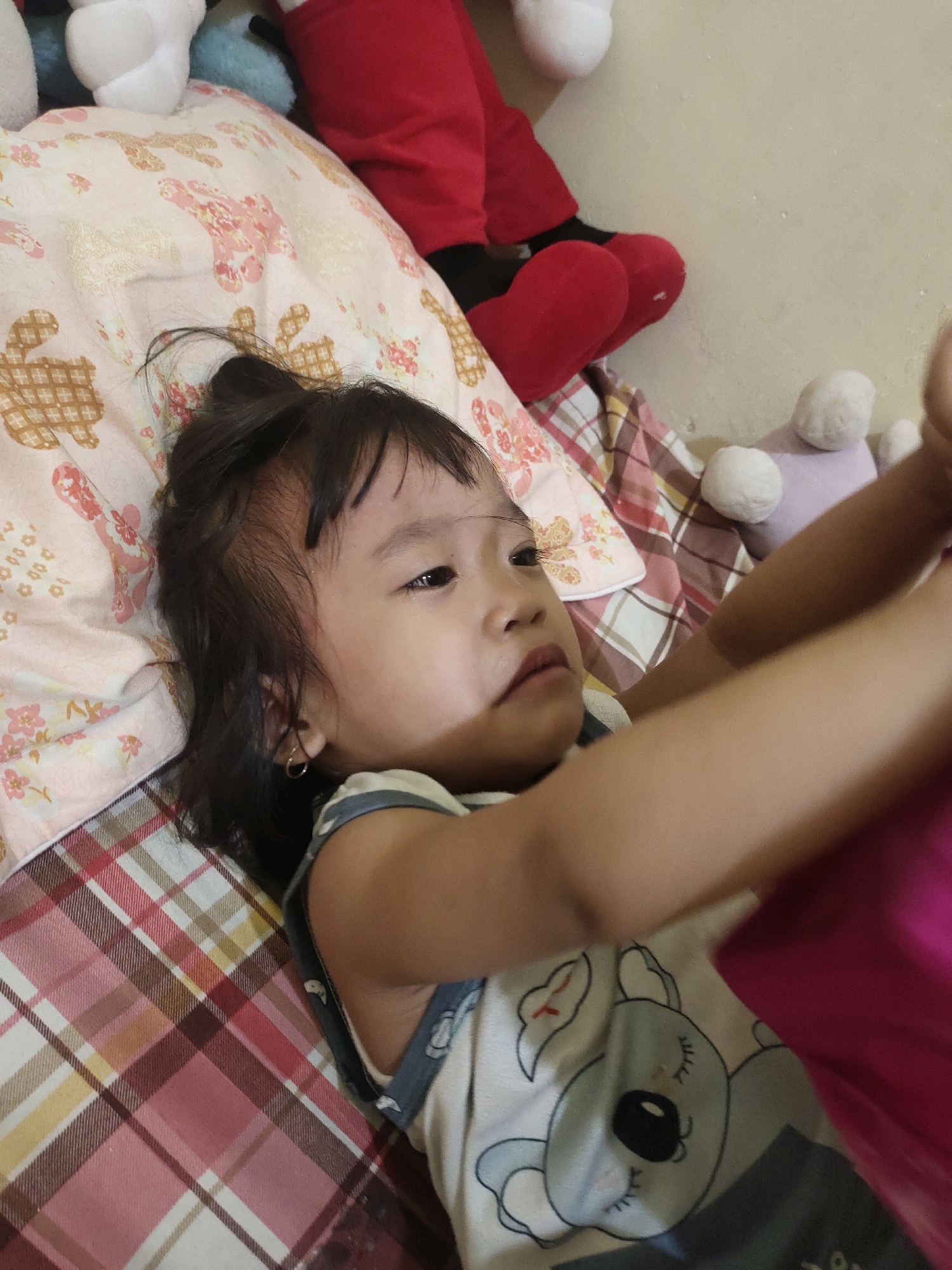 A young girl with dark hair is lying on her back on a bed. She is wearing a light-colored sleeveless shirt with a cartoon koala print. The bedspread features a pink and white floral pattern and a checkered pattern. Several stuffed animals are visible in the background, including a red and black stuffed character and white and light purple spherical stuffed toys. The girl's arms are raised, and she appears to be looking upwards. The environment suggests a domestic setting, likely a bedroom. The image was taken in Kroman, Indonesia.