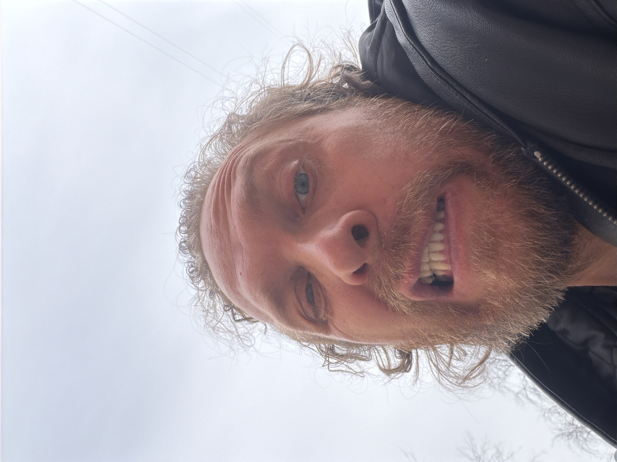 A close-up, skewed perspective selfie captures a man with curly brown hair, a reddish-brown beard, and light-colored eyes, wearing a black leather jacket. His expression shows his teeth. The background is a light gray, overcast sky with indistinct silhouettes of bare tree branches. The location is unidentifiable from the image and cannot be attributed to Saint Petersburg, Russia.