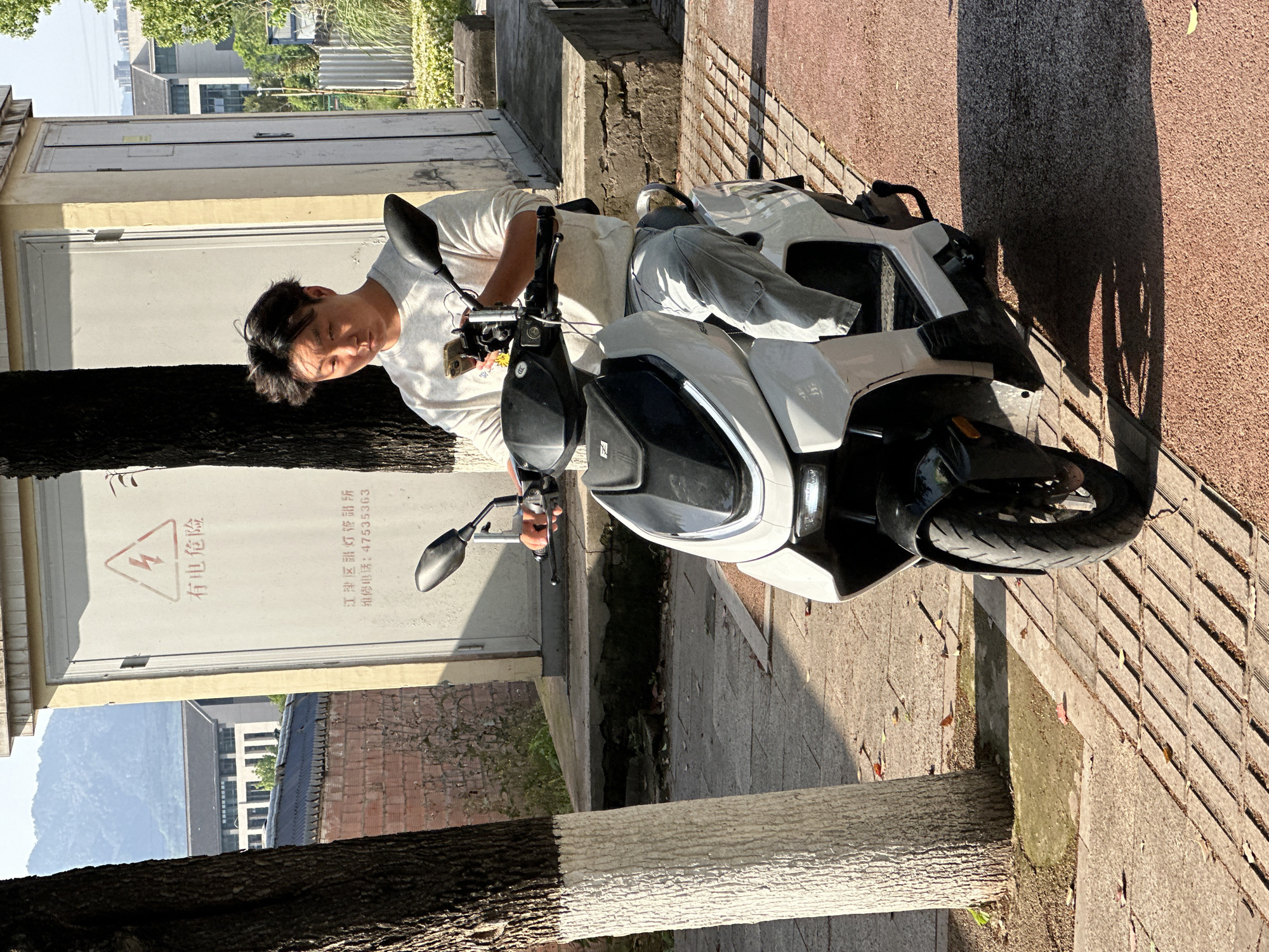 A young man is depicted sitting on a white electric scooter on a sunny day in Jiangjin, China. He wears a light-colored t-shirt and has dark hair, with his eyes appearing somewhat squinted, possibly due to the bright sunlight. He is holding a smartphone in his left hand, which rests on the scooter's handlebars.
The scooter, primarily white with black details, is parked on a grey paved sidewalk next to a reddish, textured pathway. Distinct long shadows are cast across the ground, indicating the sun is relatively low, suggesting either morning or late afternoon. Two trees are visible, one in the foreground with its lower trunk painted white.
Behind the man, there is a light-yellow utility box or shed featuring two large white doors. Prominently displayed on the right door is a red lightning bolt symbol within a triangular warning sign. Below this, Chinese characters read "有电危险" (Electric Hazard). Further text identifies the location and contact information: "江津区路灯灯管所" (Jiangjin District Street Light Management Office) and "维修电话: 47535363" (Maintenance phone: 47535363).
In the background, various buildings are visible, including one with multiple windows and another with a brick-like facade. Distant mountains rise against the sky, providing a scenic backdrop to the urban setting. The man's expression is neutral, possibly concentrating on his phone or simply resting. The scene captures a typical moment in a residential or commercial area.
