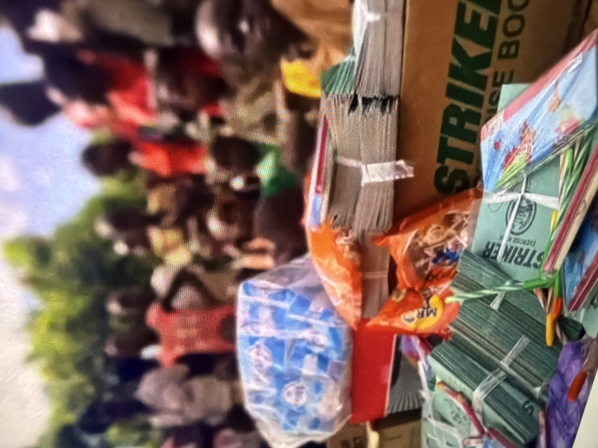 In an outdoor setting, a group of blurred people, mostly children, are gathered around tables laden with what appear to be school supplies and snacks. The foreground is dominated by stacks of notebooks, some wrapped in green paper with "STRIKER" visible on them, others with colorful cartoon-like designs. There are also plastic bags filled with blue items, possibly soap or other small goods, and orange bags that may contain snacks. A large cardboard box with "STRIKER" printed in green lettering is visible behind the supplies. The background is a blur of greenery and sky, suggesting an environment with natural elements. The lighting is bright, indicating it is daytime.