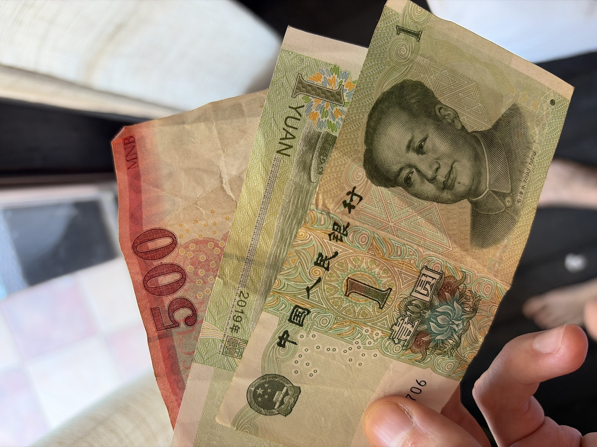 A hand holds three banknotes against a softly blurred, light-colored background, suggesting an indoor setting. The visible money includes two Chinese 1 Yuan banknotes and one Mongolian 500 Tugrik banknote.
The two green 1 Yuan notes prominently feature a portrait of Mao Zedong. Text visible on these notes includes "1 YUAN", the numeral "1", the Chinese characters "中国人民银行" (People's Bank of China), and the dates "1893-1976" (Mao Zedong's birth and death years). One of these notes also clearly displays "2019年", indicating its issue year.
The third banknote, partially obscured and appearing reddish-brown, shows the large denomination "500". The letters "MNB" are visible along its edge, identifying it as a 500 Mongolian Tugrik note, likely issued by the Mongolian National Bank.
The activity shown is a person holding these different denominations of foreign currency. The photograph was taken in Budva, Montenegro.