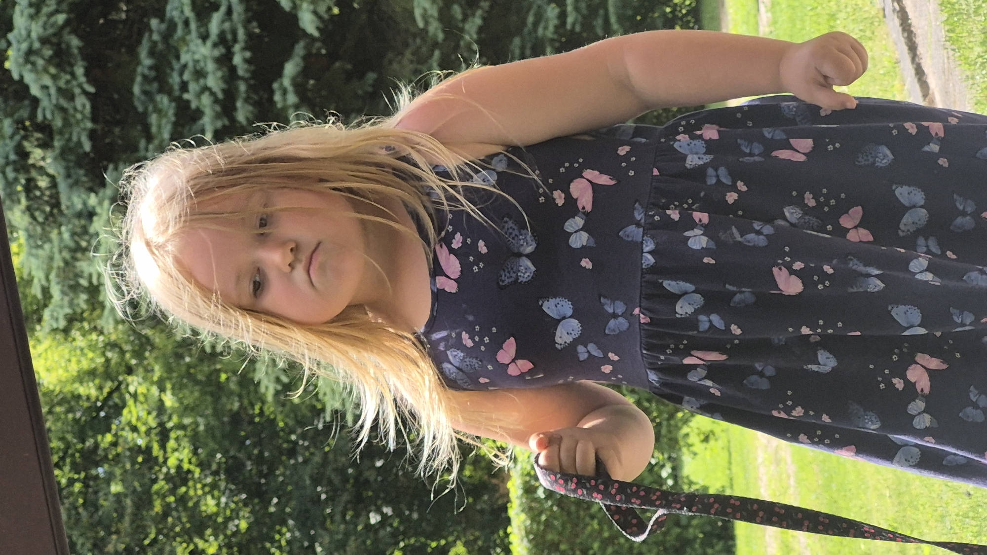 A young girl with blonde hair stands outdoors, holding a dark-colored strap with a pink and black pattern. She is wearing a dark blue dress adorned with pink and blue butterflies. Her hair is windswept, and she has a pouting expression. The background consists of lush green trees and grass. The image appears to have been taken in a park or a garden. It is not possible to determine the exact location within Švenčionys, Lithuania, from the provided image.