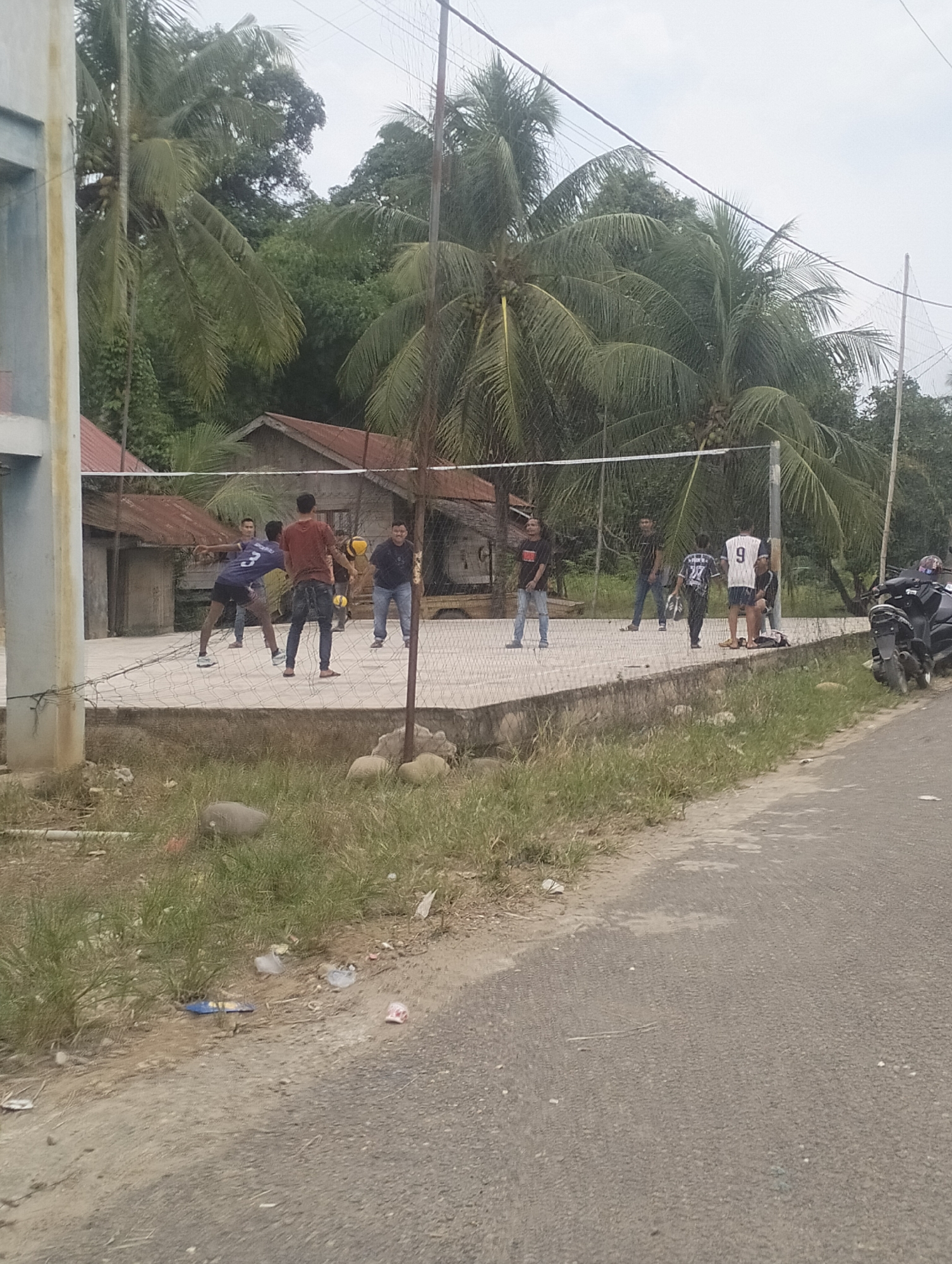 Seven young men are playing volleyball on an outdoor court. The court is made of concrete and has a net stretched across the middle. Palm trees and tropical foliage surround the court, and two small wooden buildings with tin roofs are visible in the background. A paved road runs along the right side of the image, with a motorcycle parked nearby. The sky is overcast.