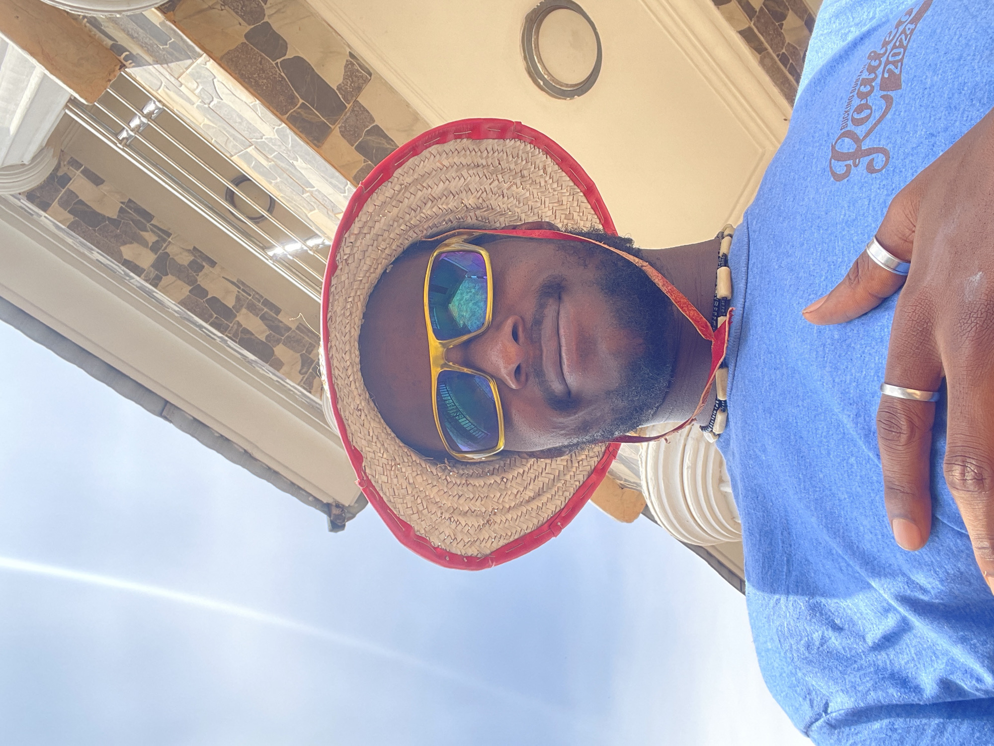 A dark-skinned man with a beard is captured in a low-angle, close-up shot, looking downwards toward the camera with a relaxed, slight smile. He is wearing a straw hat with a red brim, yellow-framed sunglasses with reflective blue/green lenses, and a beaded necklace featuring black and light-colored beads. His light blue t-shirt is visible, and on his right hand, two silver-colored rings are worn: one on the index finger and one on the ring finger.
The setting appears to be outdoors or under a covered outdoor area, likely a porch or veranda. Above and to the left of the man, a bright blue sky is visible, with a sunbeam or lens flare stretching across it, indicating clear, sunny daytime conditions. To the man's right, a distinct ceiling or overhang structure is seen, featuring decorative geometric tiles in shades of brown, beige, and grey. A circular recessed light fixture is embedded in this ceiling. A light-colored, possibly white, architectural pillar or detail is also visible on this side.
On the man's light blue t-shirt, the text "Rockfest 2024" is clearly legible, along with partially obscured words above it that appear to read "BUCKING BOARS." The overall mood is casual and relaxed.