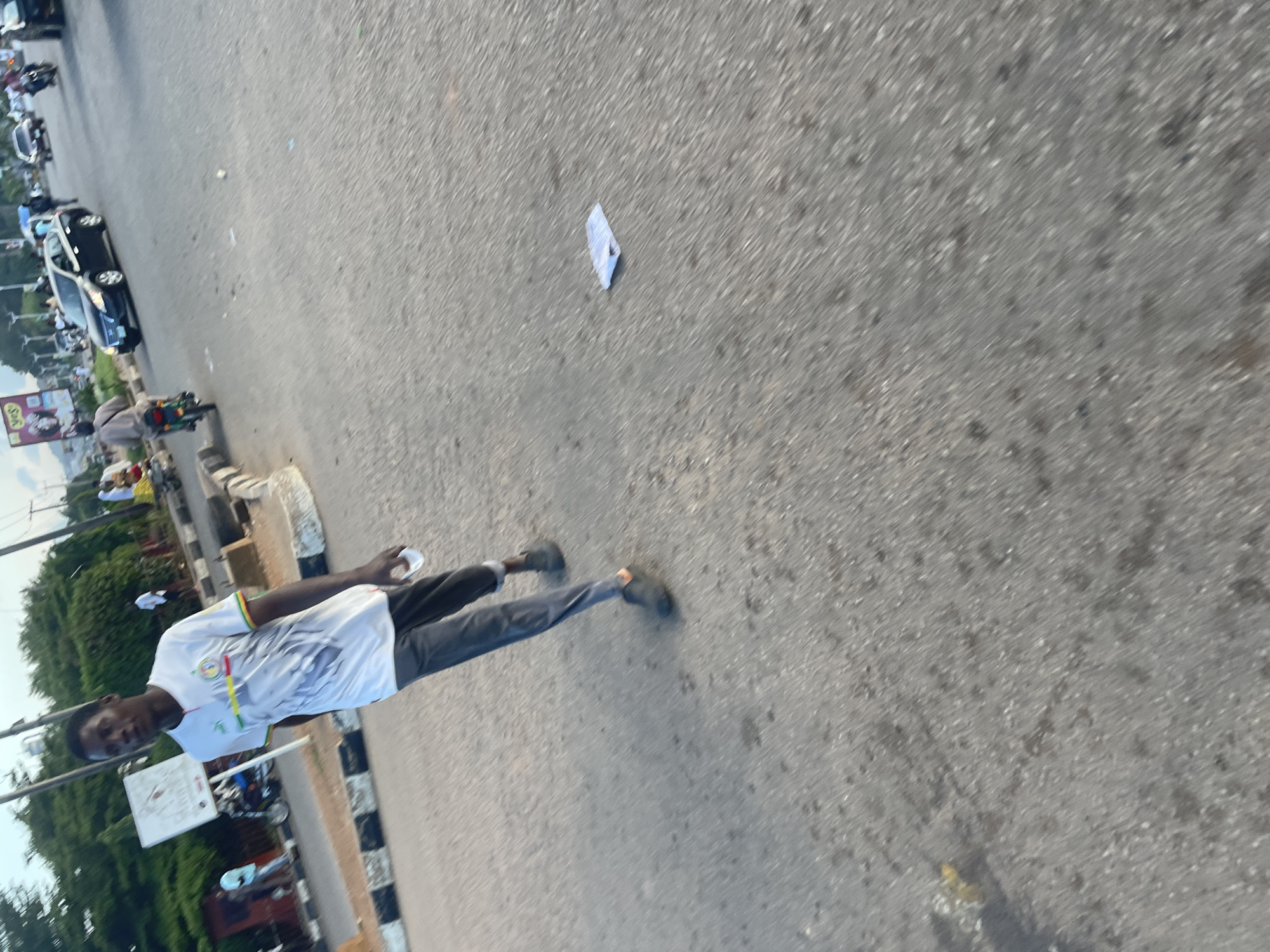 In Jos, Nigeria, on a clear day, a young man walks across a paved road. He is wearing a white t-shirt with green, yellow, and red accents on the collar and sleeves, and a graphic design on the front. He also wears dark gray pants and dark shoes. He holds a white object in his right hand. The road has a striped curb on the left, and vehicles, including a dark sedan, motorcycles, and other cars, are visible in the background, suggesting it is a busy street. The scene appears to be captured from a low angle, with trees and a signpost visible on the left side of the frame.