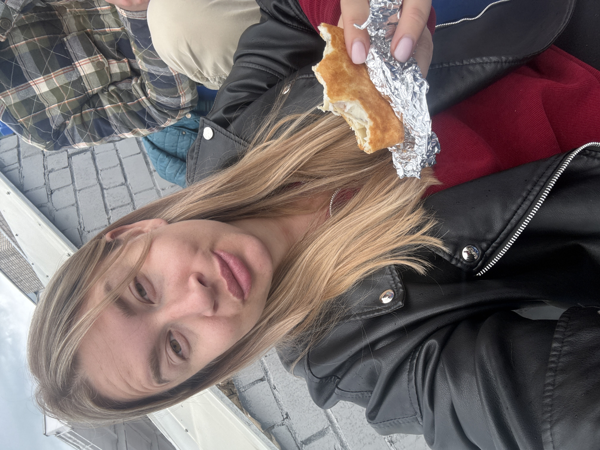 A blonde woman wearing a black leather jacket and a red top is holding a partially eaten sandwich wrapped in aluminum foil. Another person in a plaid shirt and beige pants sits beside her. The setting appears to be outdoors, potentially on a boat, with brick-patterned ground visible. The location is Veliky Novgorod, Russia.