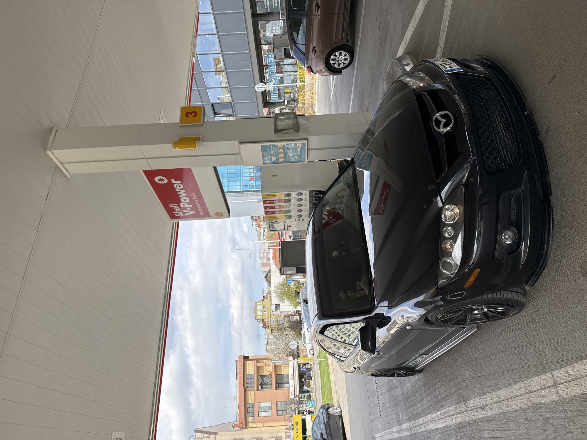 The image captures a black Mazda car parked at a Shell gas station. The car is positioned at a gas pump numbered '3'. In the background, there is an array of buildings, suggesting an urban setting. There is a sign that says NITH STIGER. The buildings are located in the city of Sofia, Bulgaria.