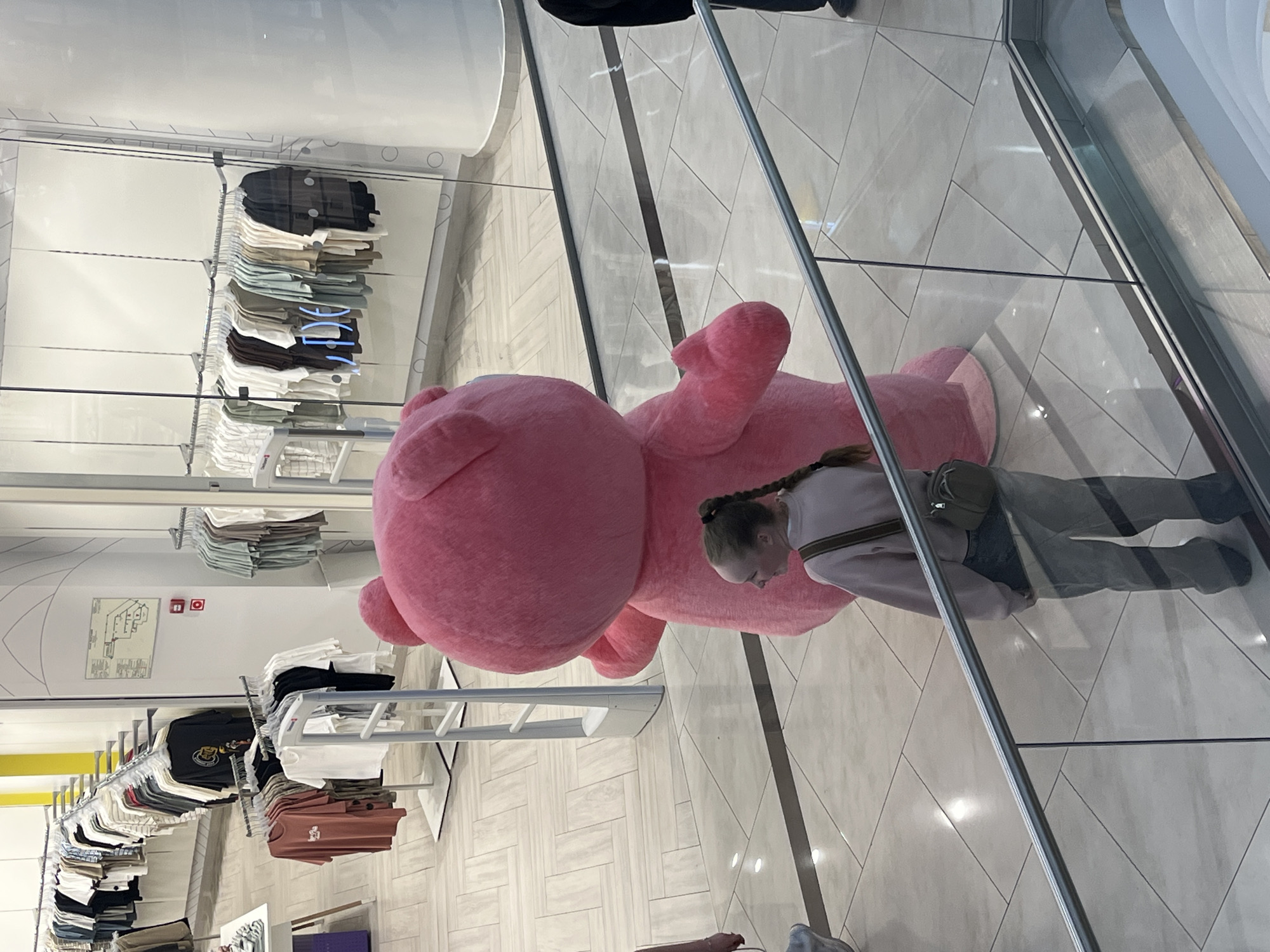 This image captures an indoor scene within a modern clothing retail store, located in Perm, Russia. A large, bright pink mascot costume, resembling a bear, stands with its back to the viewer in the middle-ground, positioned near white electronic article surveillance (EAS) security gates at a store entrance. In the foreground, viewed from an elevated perspective through a glass and metal railing, a woman with light hair styled in a braid is observed from behind, her head turned slightly to the right as she looks towards the pink mascot. She is dressed in a light-colored long-sleeved top, light-toned jeans, and carries a small, olive-green cross-body bag. On the far right edge of the frame, a partial view of a person's lower leg and foot, wearing dark clothing and footwear, is visible behind the railing. The retail environment features multiple clothing racks displaying various t-shirts and casual wear in a range of colors, including neutrals, earth tones, and muted pastels. A floor plan diagram and two red emergency call buttons are mounted on a white wall to the left of the store entrance. The floor consists of light-colored rectangular tiles arranged in patterns, and the entire area is brightly illuminated by overhead lighting, which reflects off the tiled surface and glass panels.