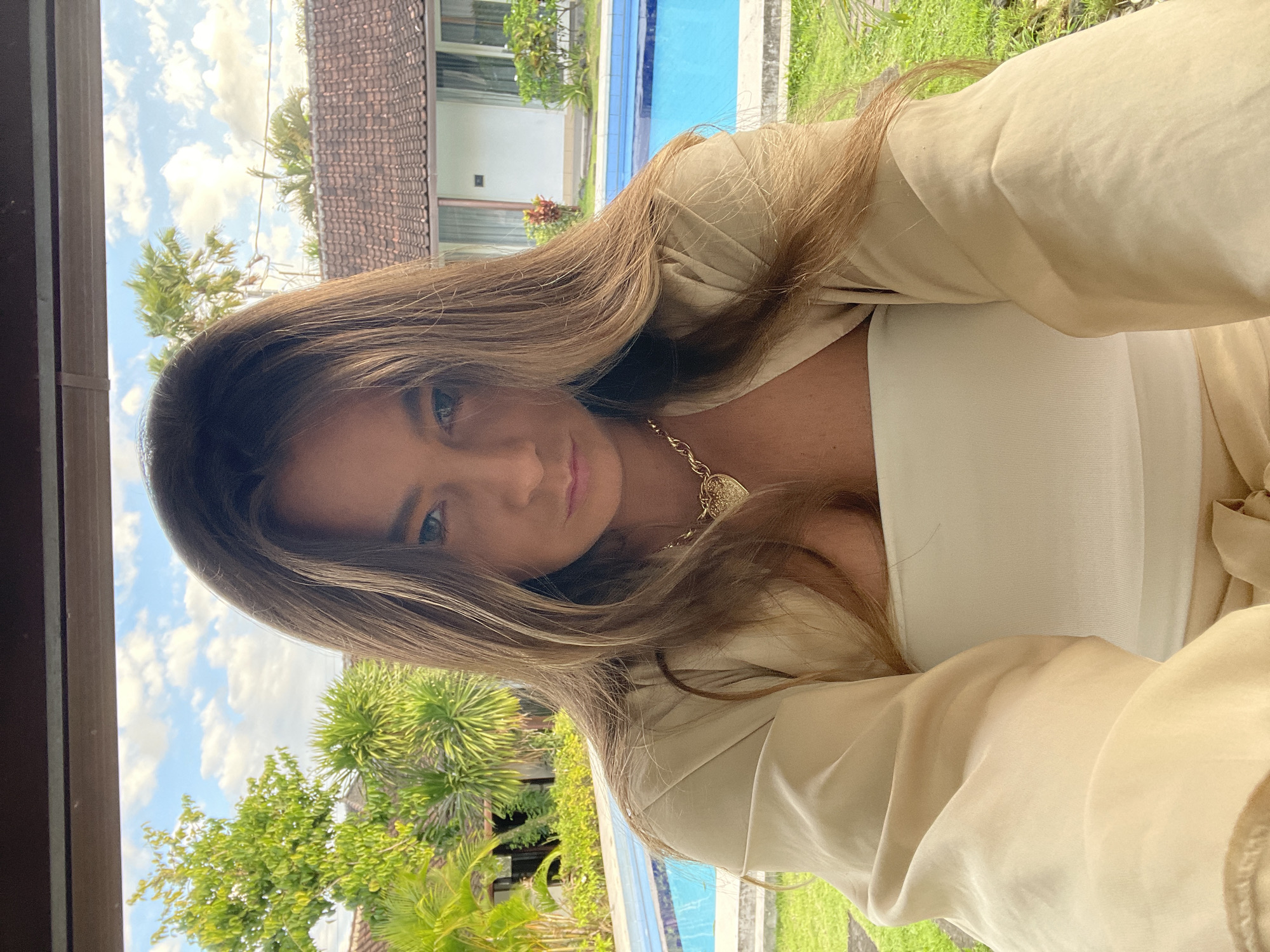 A woman with long, blonde-brown hair and a serious expression is taking a selfie outdoors. She is wearing a beige off-the-shoulder top over a white strapless top and a gold necklace with a heart-shaped pendant. Behind her is a swimming pool with blue water, surrounded by green grass and lush tropical foliage. In the background, there are buildings with red-tiled roofs and palm trees. The scene is set in North Kuta, Indonesia. The sky is blue with scattered white clouds.