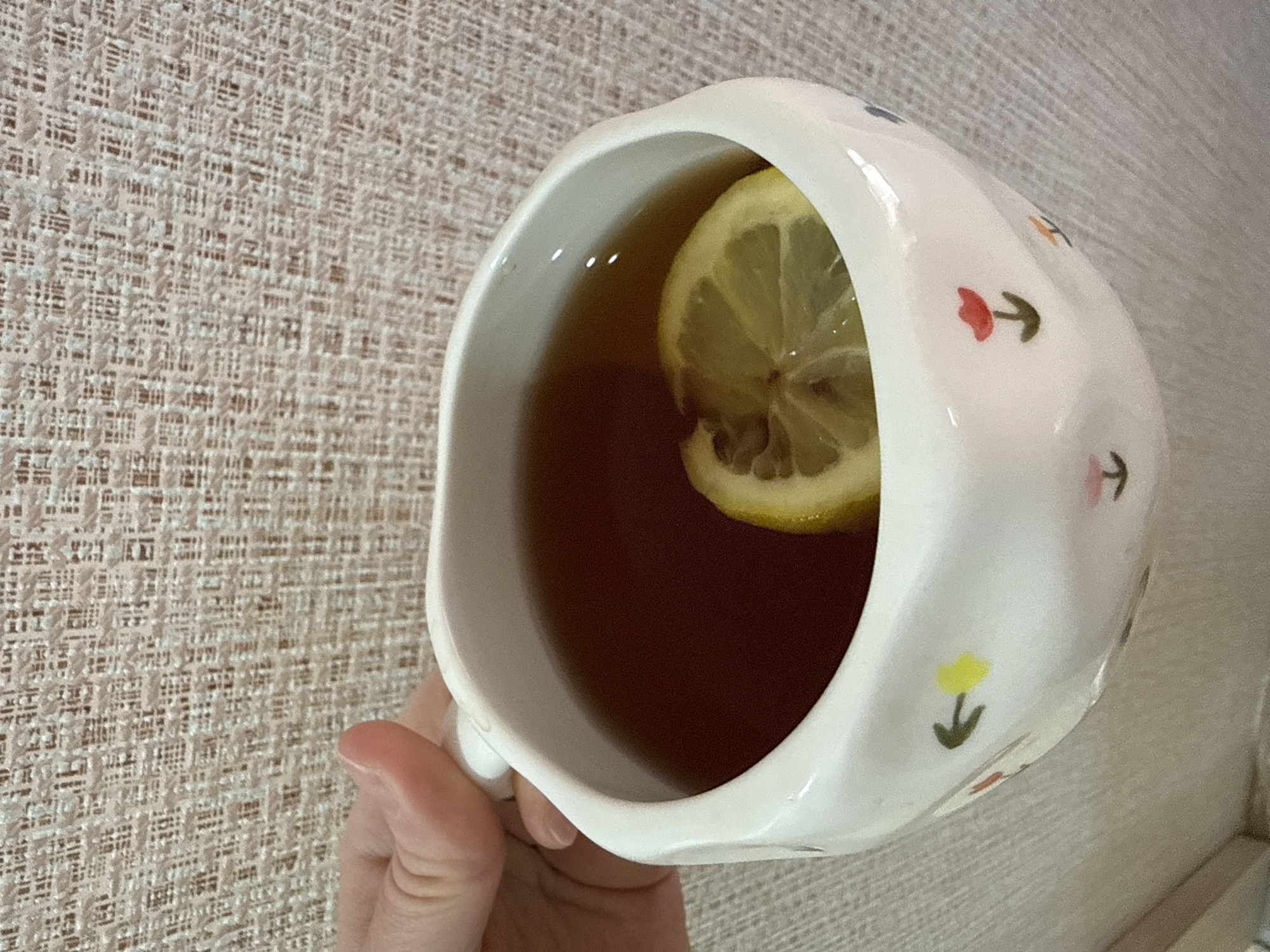 A close-up, high-angle shot captures a person's hand holding a white ceramic mug. The mug contains a dark brown liquid, likely tea, with a single slice of fresh yellow lemon floating on top. The mug itself is adorned with small, simple, and colorful floral patterns, resembling tulips, in shades of red, yellow, orange, and blue, with green stems. The hand is visible gripping the mug's handle from the left side of the frame. The background is a textured, light beige or off-white fabric, possibly a blanket, tablecloth, or upholstered surface, featuring a fine, woven, grid-like pattern. The lighting suggests an indoor setting, but the time of day, specific location, and emotions are not discernible. No text is visible in the image.