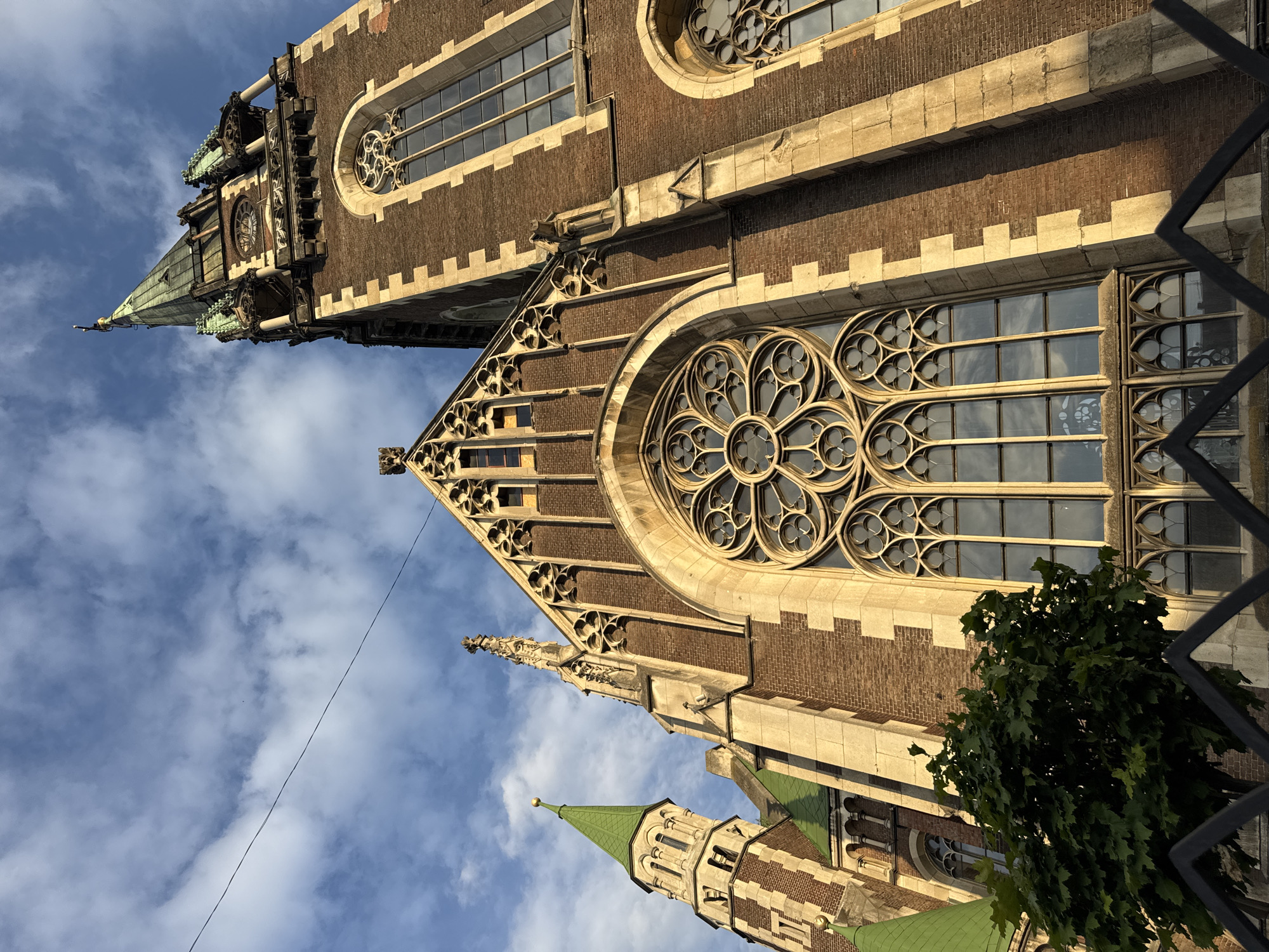 The image shows a low-angle, tilted view of the exterior of a large, ornate brick building with Gothic architectural features. The building's facade features several large, arched windows with intricate tracery, including a prominent rose window. The stonework surrounding the windows is light-colored and forms a decorative pattern along the top of the building. A tall, spired tower with a green roof and decorative elements is visible on the left side, partially obscured by the main structure. Another smaller green-roofed tower can be seen in the lower right of the frame. The sky is blue with scattered white clouds, suggesting daytime. A dark metal fence is visible in the foreground on the right, and a dark wire stretches diagonally across the sky. A lush green tree with dense foliage partially obscures the lower part of the building on the right. The building's brickwork and elaborate detailing suggest it is a historic religious or public structure, likely a cathedral or church. The style is consistent with Gothic architecture. There is no visible text in the image. The overall impression is of grandeur and historical significance. The lighting suggests it is either early morning or late afternoon.