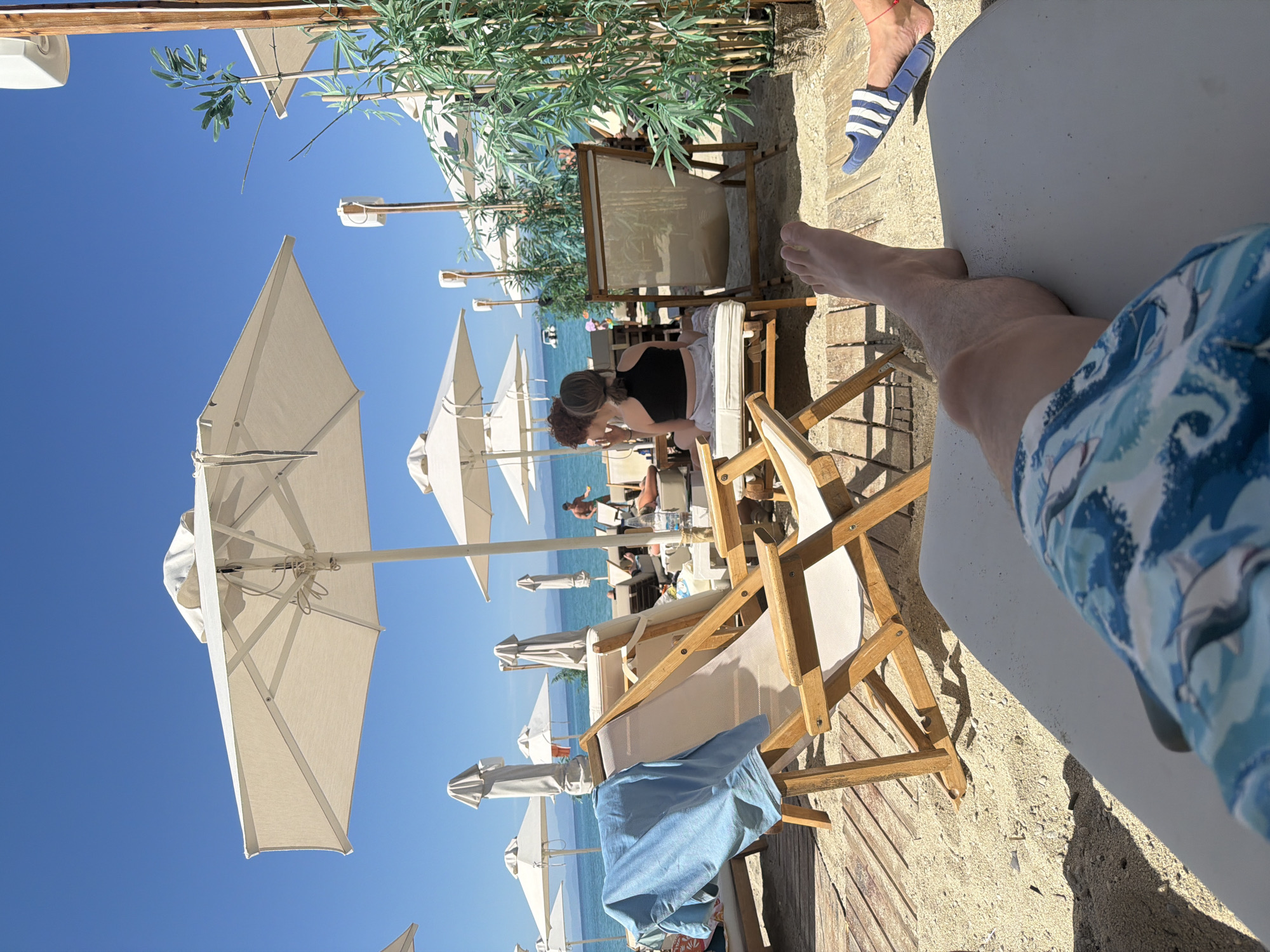 The image shows a beach scene in the Pallini Municipal Unit, Greece. The viewpoint is from a person reclining on a white lounge chair, wearing blue patterned swim trunks. The right leg and foot of the person are visible. To the left and further away are other lounge chairs, some occupied by people. Some are under large white parasols. Behind the chairs is a wooden deck leading down to a sandy beach and the Aegean Sea. In the background, other people are visible and there's light vegetation above some of the loungers. The sky is clear and blue. Another leg and a foot with blue and white striped sandals can be seen in the upper right corner of the image.