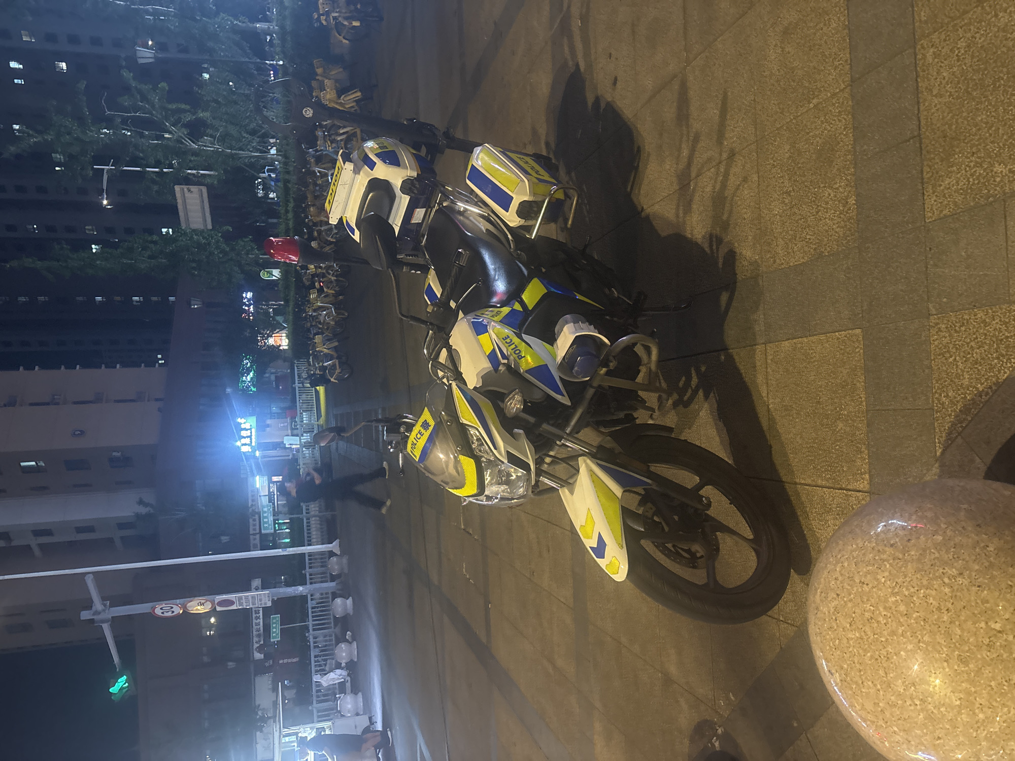 The image shows a police motorcycle parked on a tiled walkway at night. The motorcycle is white and blue with yellow and blue checkered markings and the word "POLICE" visible on several parts. It has storage compartments on the back and sides, and a red flashing light on top. In the background, there are tall buildings with lit windows, streetlights, trees, and numerous yellow shared bicycles. A person is walking in the background to the left. Traffic lights are visible on a pole, with a green light illuminated. A speed limit sign (30) is also present on the pole. The scene appears to be in a public area in the city of Suzhou Industrial Park, China, given the presence of shared bicycles and the typical urban night setting.
