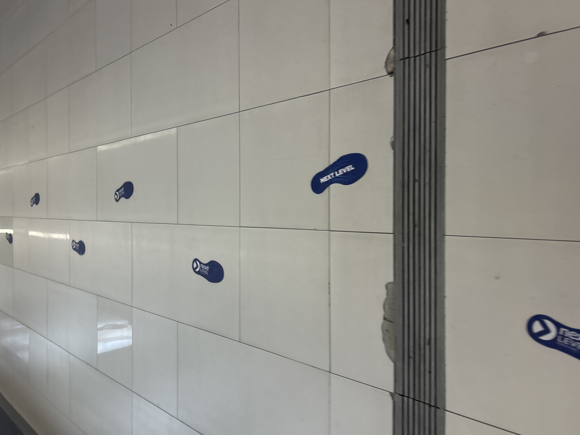 The image shows a tiled wall in an indoor location, most likely a passageway. There are several blue foot-shaped stickers on the wall, each bearing the words "NEXT LEVEL." The stickers are spaced out along the wall, suggesting they are guiding pedestrians. A gray textured strip runs vertically on the wall. Given the presence of directional stickers, this may be a walkway in the city of Plovdiv, Bulgaria.
