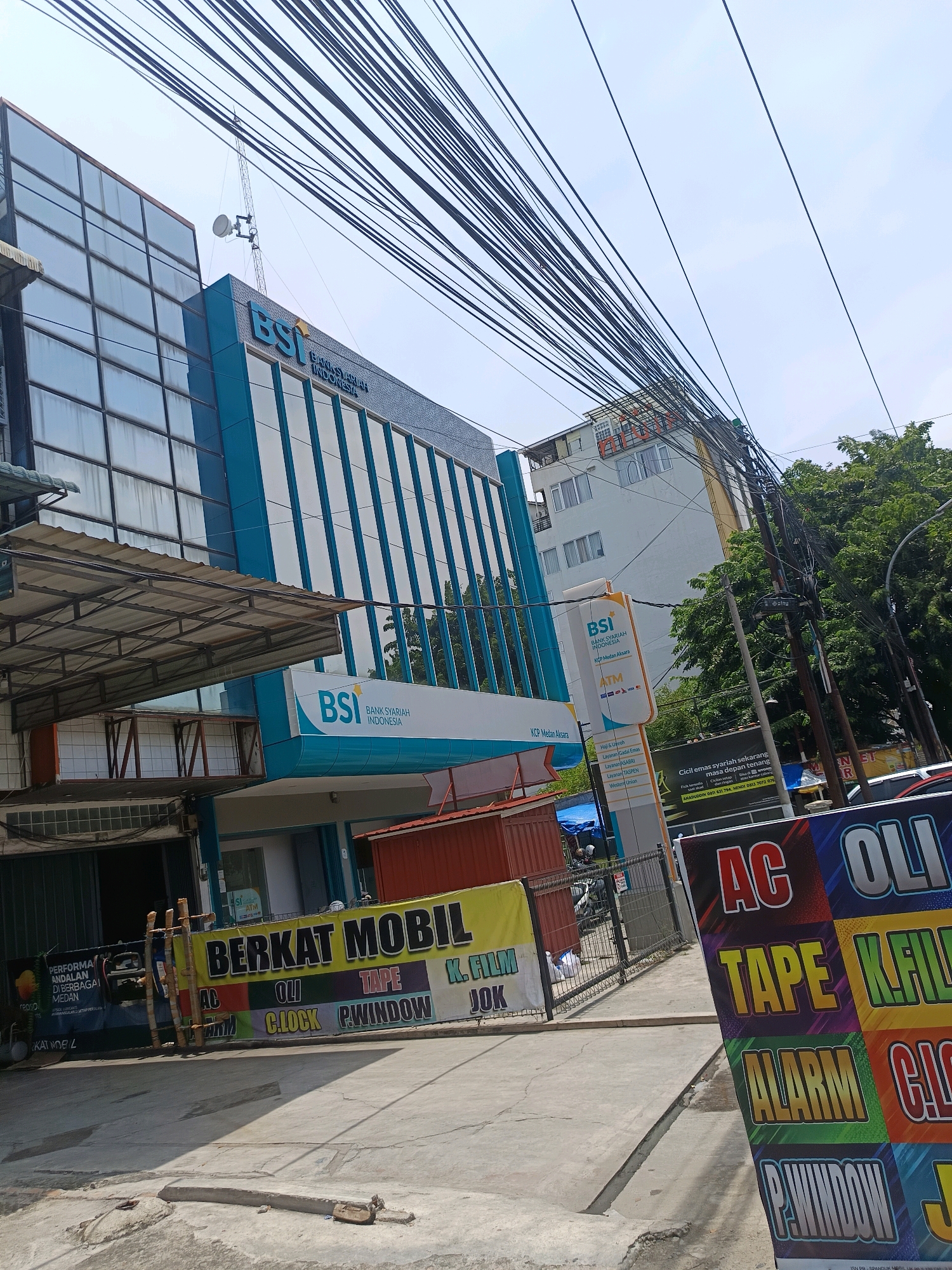 The image shows a street view in Percut Sei Tuan, Indonesia, featuring a prominent Bank Syariah Indonesia (BSI) building. The BSI building is modern, with a blue and white facade and large glass windows. To the left of the BSI building is another building with a similar glass facade. In front of the BSI building, a street-level commercial space is visible, with signage indicating services like "BERKAT MOBIL" (Car Blessing), "OLI" (Oil), "TAPE", "K. FILM", "ALARM", and "P. WINDOW". There is a street with parked vehicles and some trees in the background. Overhead, a dense network of power lines stretches across the sky. The overall scene depicts a commercial area on a clear day.