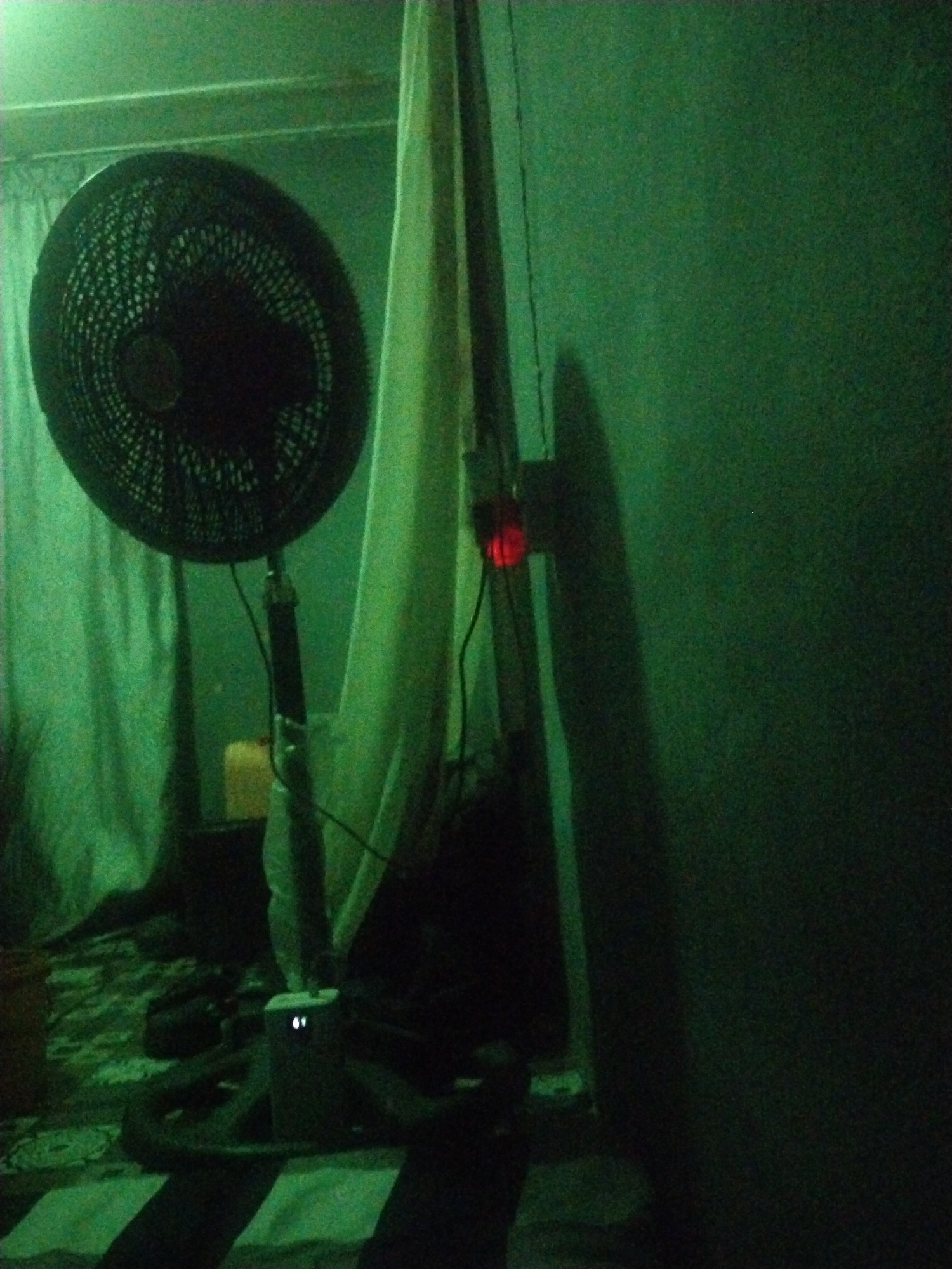 The image depicts a dark room in a location identified only as Unknown, Unknown. An electric fan with a black grill stands prominently, positioned near a light-colored curtain. A power strip with a red light is plugged into the wall. The room's walls are a dark shade of green. The floor has a striped pattern. Other objects are present, but obscured by the low lighting conditions. There are no people visible in the image.