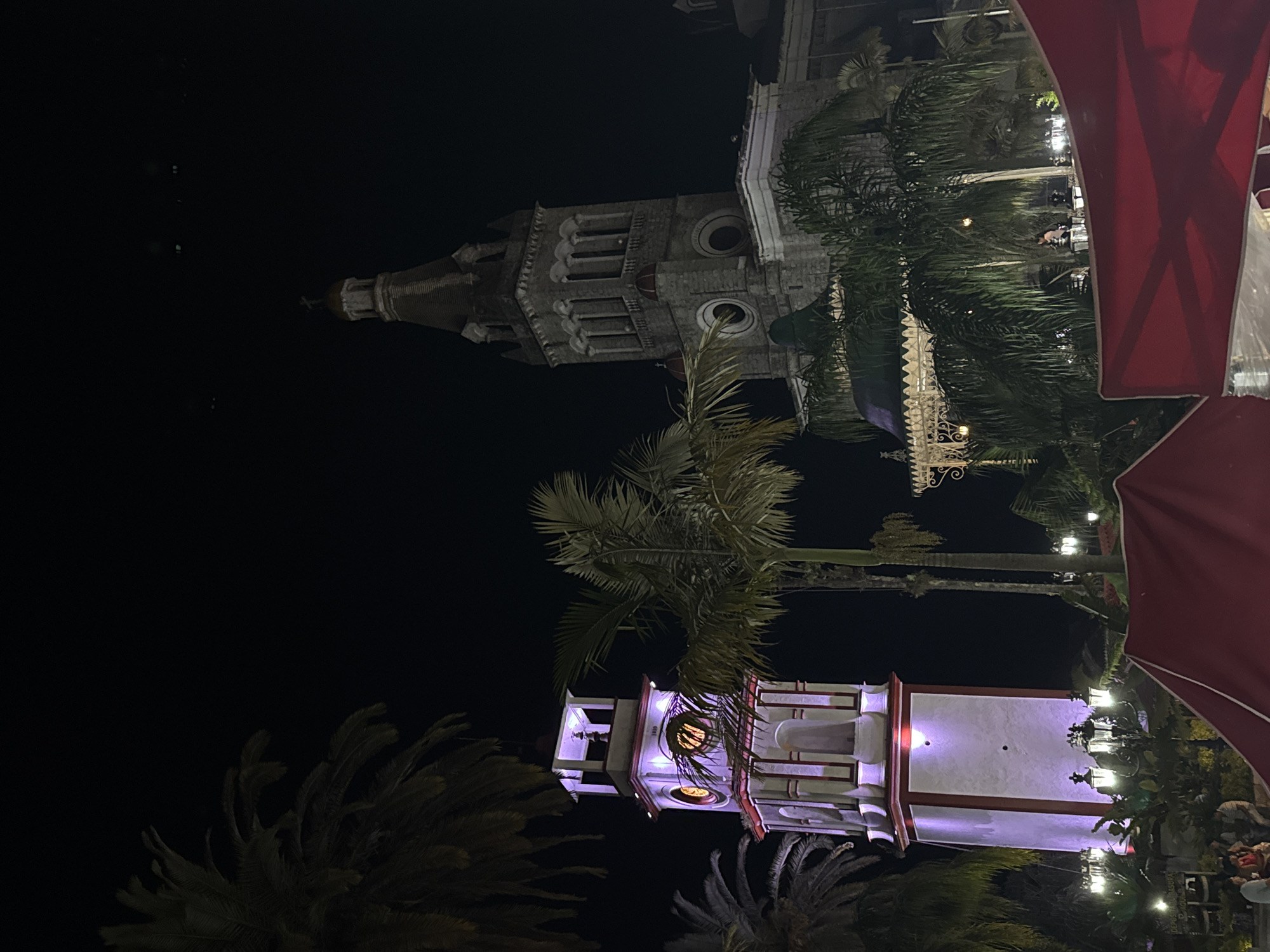 The image captures a night scene in Cuetzalan, Mexico, showcasing two prominent architectural structures. The first is a gray stone church tower with a steeple, featuring arched windows and detailed stone carvings. The second structure is a square, white tower with red trim, illuminated with purple light, revealing circular openings where a clock or design is present. Palm trees are scattered around both structures, adding a tropical ambiance to the setting. Part of a red awning is visible in the foreground, suggesting a restaurant or outdoor seating area. People are seen faintly gathered at the base of the white tower. The overall atmosphere is one of a lively and illuminated town square at night.