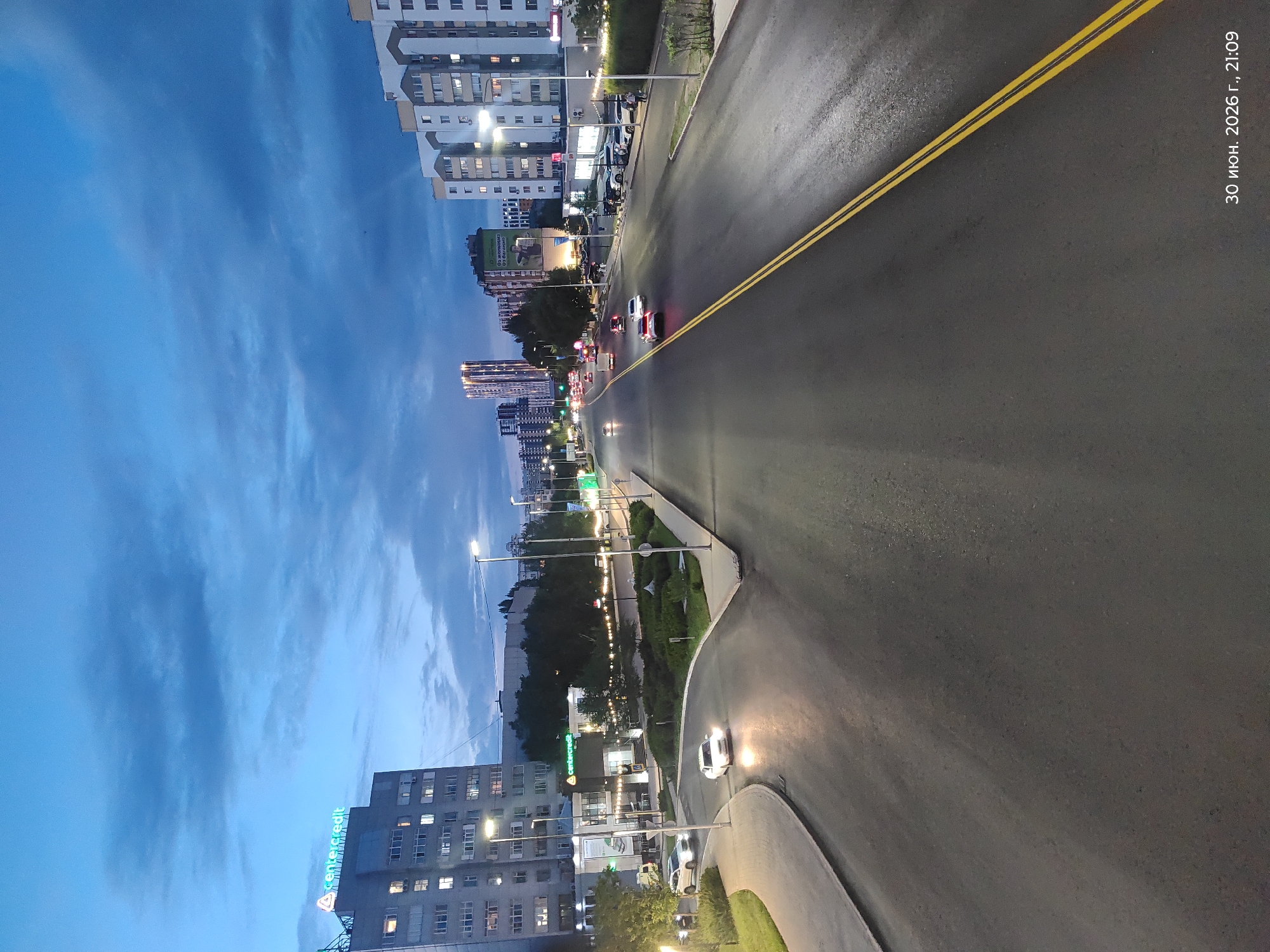 This is a photograph taken at dusk in Astana, Kazakhstan. The image captures a street scene with several cars driving on a wet asphalt road, indicated by reflections of streetlights. The sky is a deep blue, suggesting early evening or twilight. On the left side of the road, a row of modern, multi-story buildings lines the street, with some windows illuminated.
Notable details include a white car with its headlights on, and another white car further down the road. There are streetlights illuminating the sidewalk and the road. A sign with the text "centercredit" is visible on one of the buildings. Another sign in Russian, "30 июня 2026 г., 21:09", indicates the date and time the photo was taken. The presence of cars and illuminated buildings suggests an urban environment. The wet road might imply recent rain or high humidity. The overall mood is calm and urban, characteristic of a city at the end of the day.